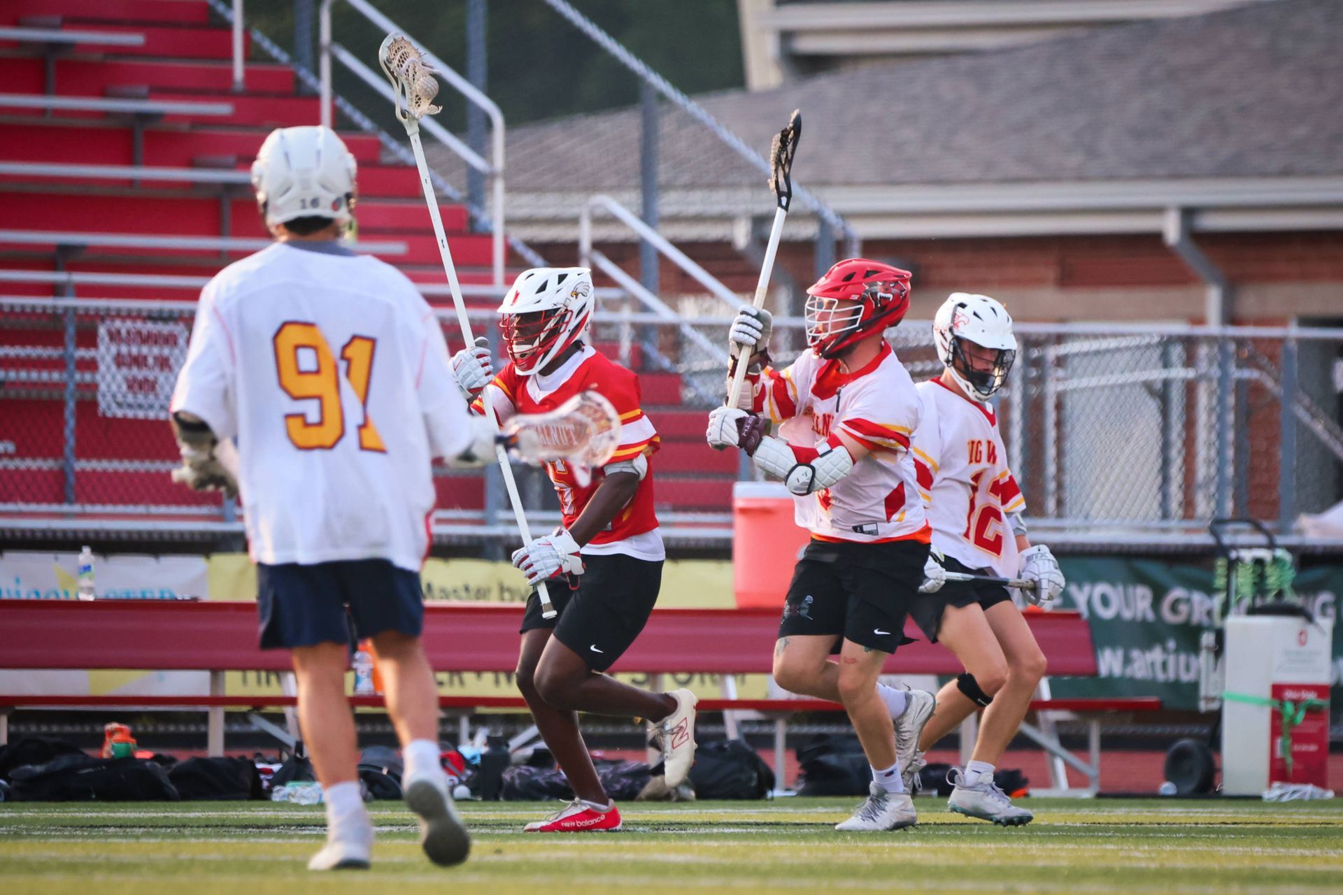 Lacrosse players in red and white sprint near the goal as an opposing player in white watches.