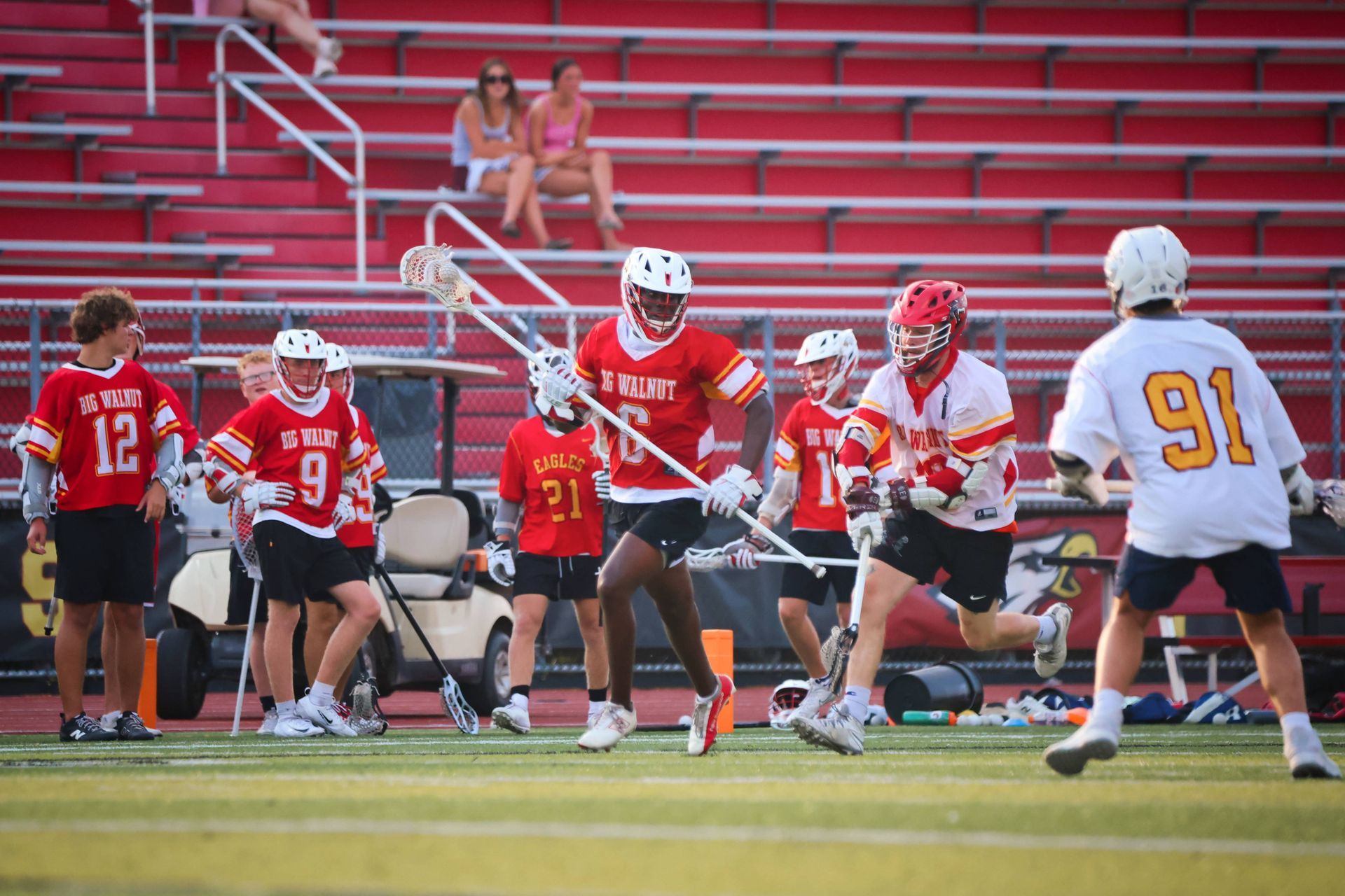 Lacrosse players in red and white jerseys compete on a field in front of red bleachers.