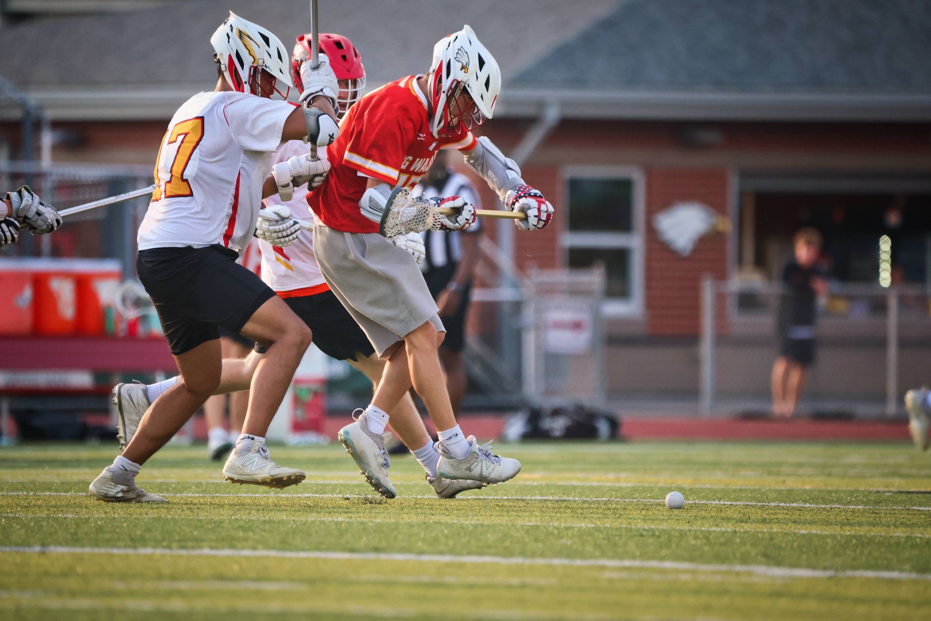 Two lacrosse players in red and white uniforms contesting a ground ball on a field