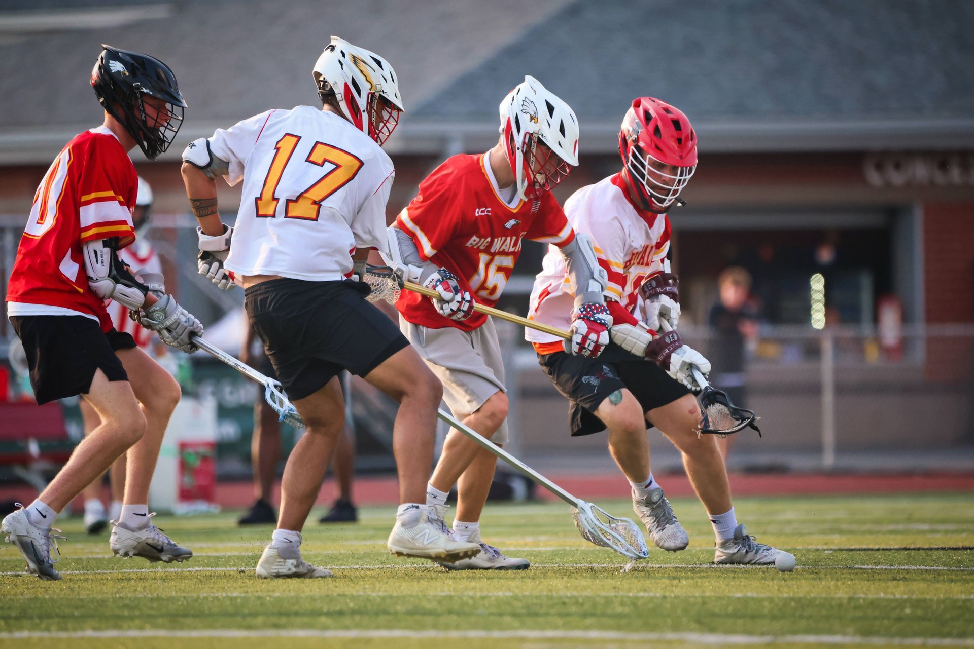Lacrosse players in red and white uniforms vie for the ball on a grass field.