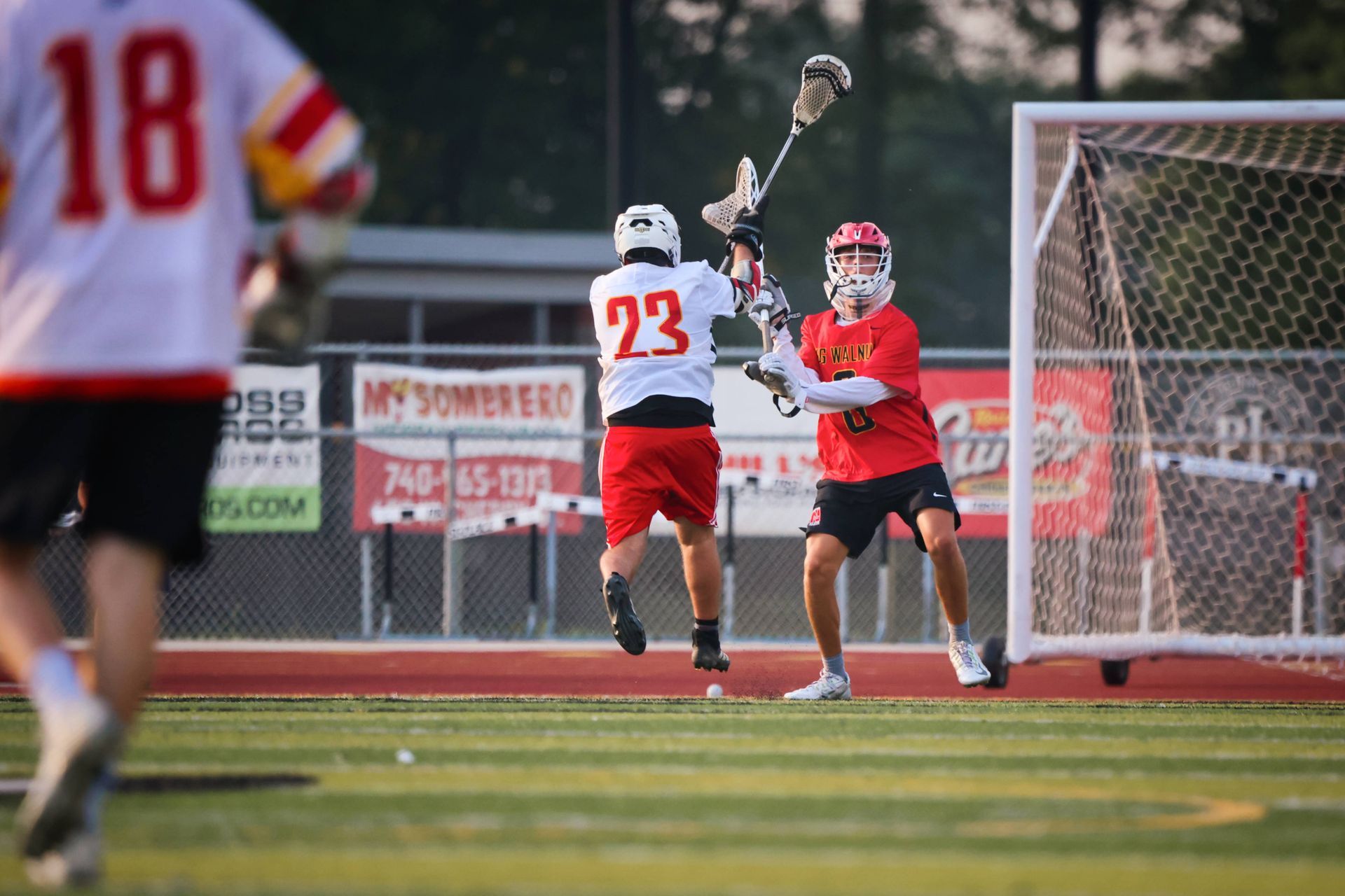 Lacrosse players in red and white battle near the goal on a field at dusk.