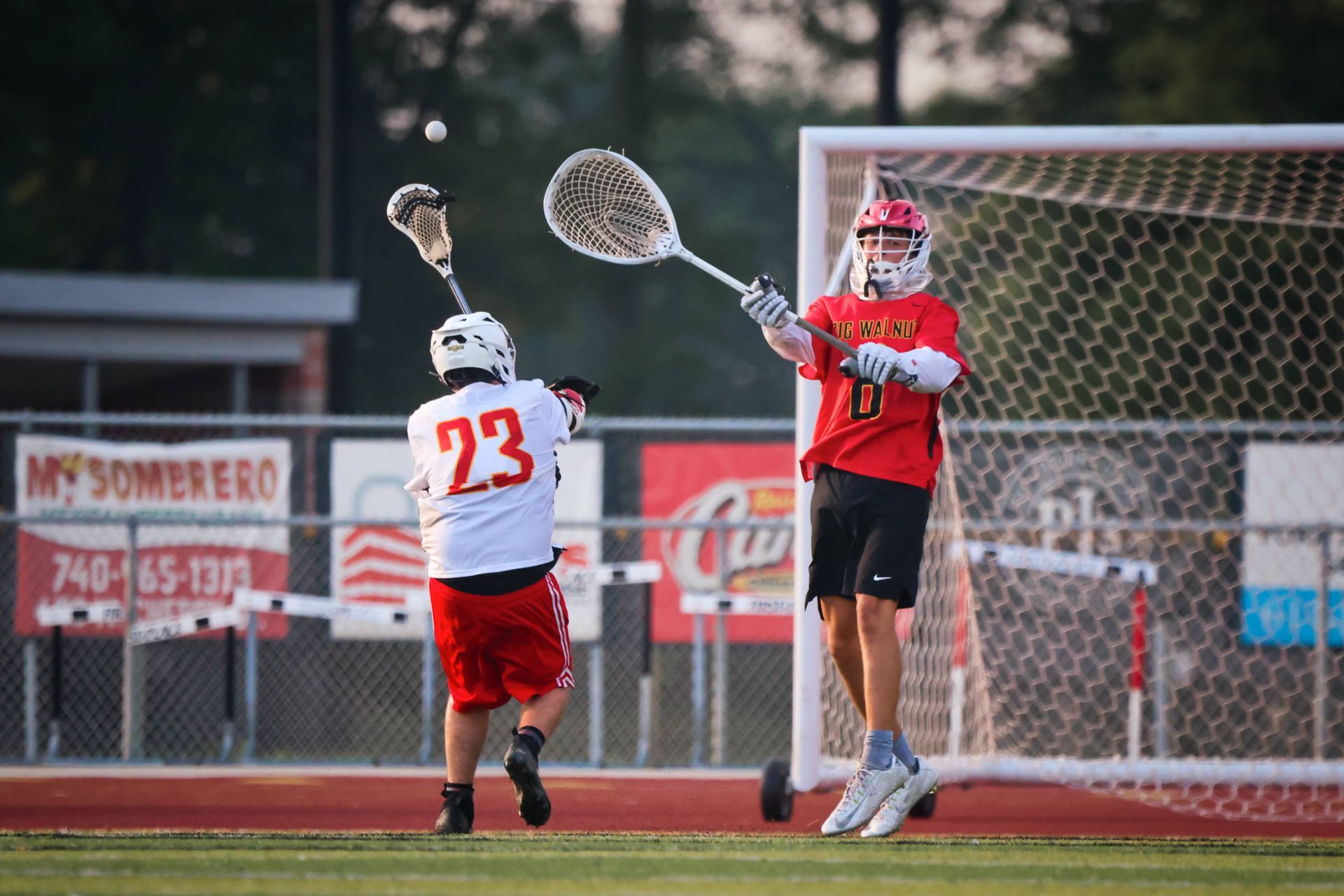 Two lacrosse players in red and white contest a shot near the goal on a field at dusk