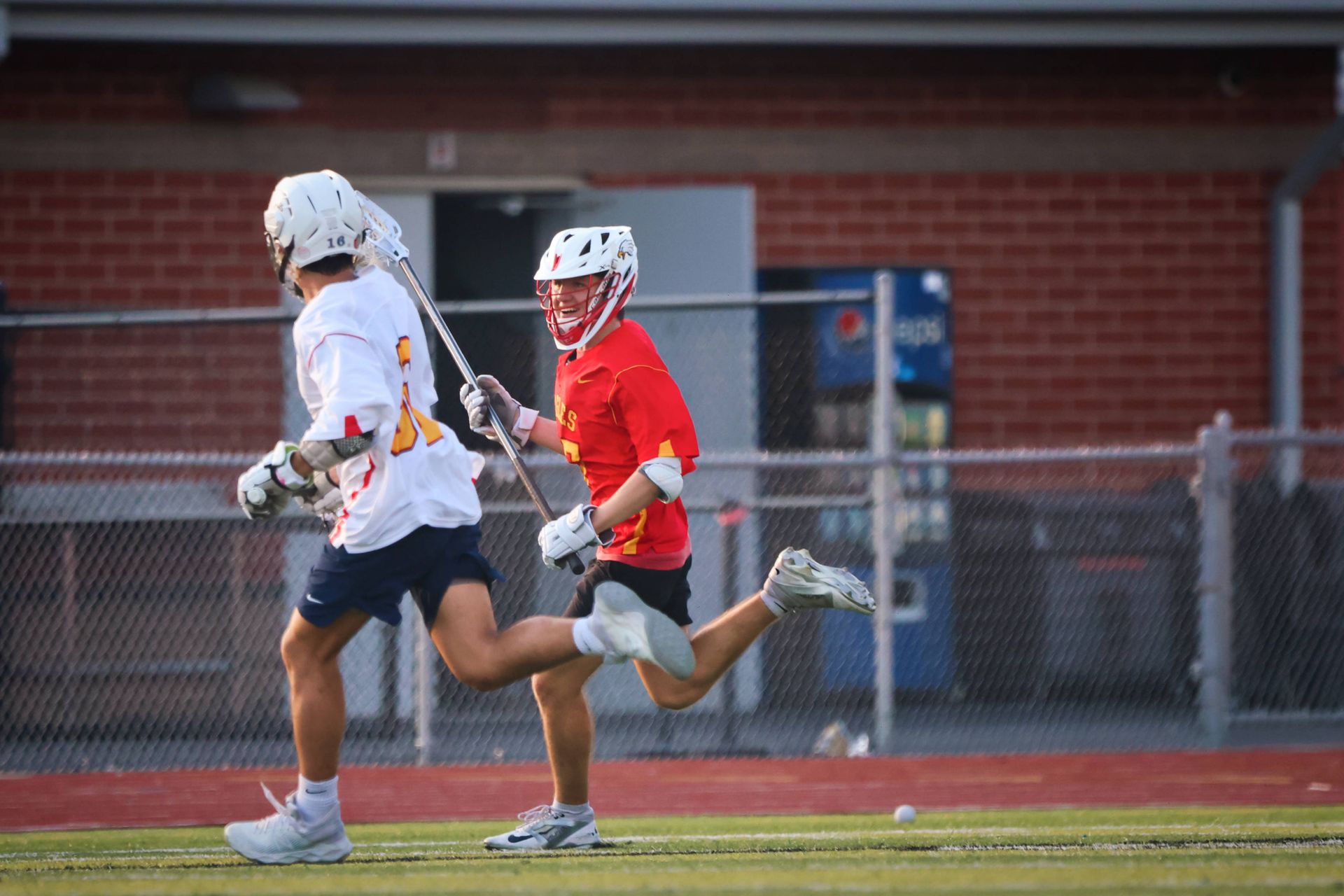 Two lacrosse players sprint on a field, one in white and one in red, near a chain-link fence.