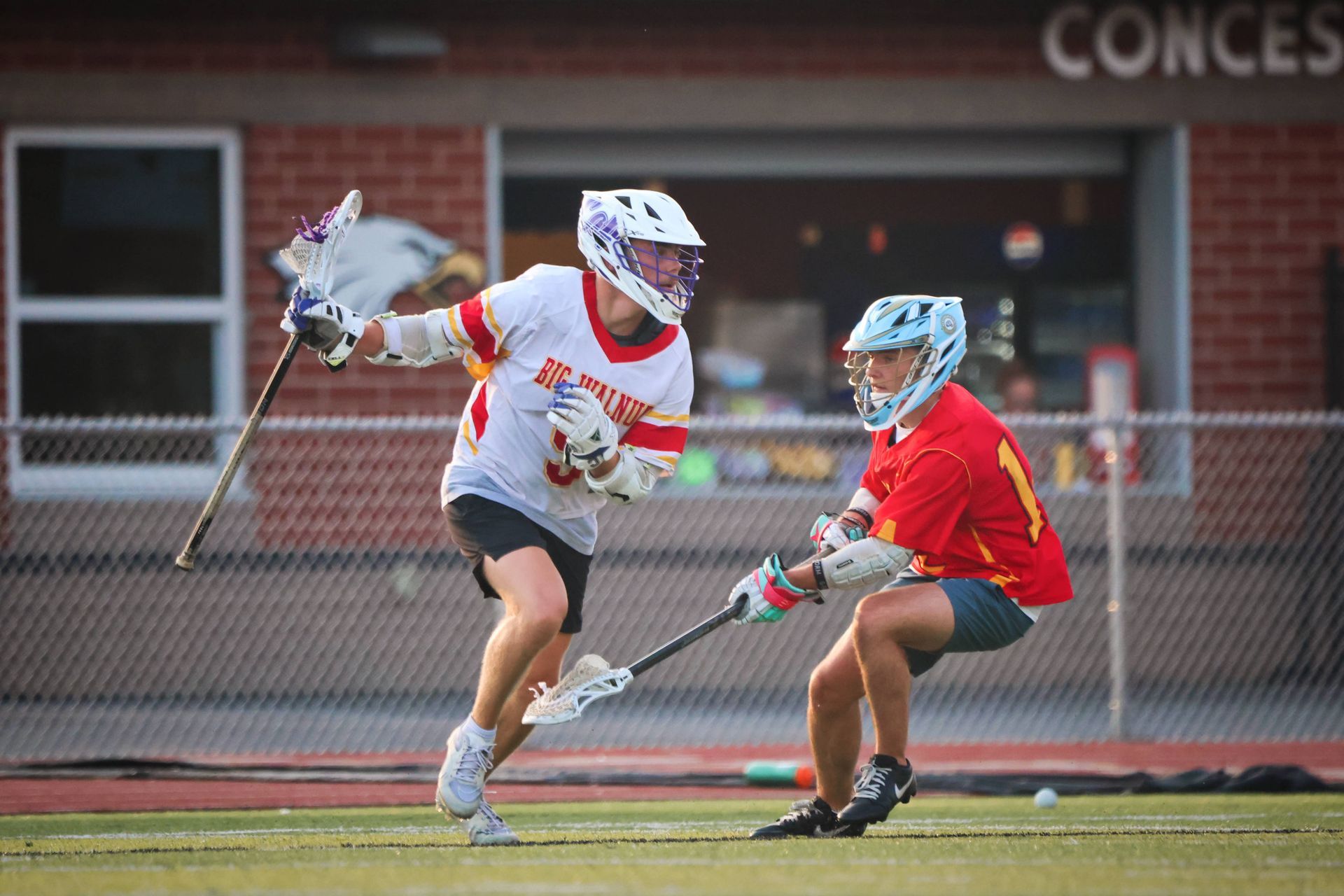 Two lacrosse players in white and red sprint and defend on a field near a brick building.