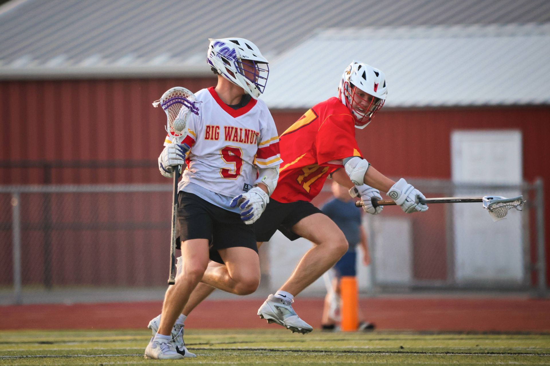 Two lacrosse players in red and white uniforms run on a field, one cradling the ball near a fence.