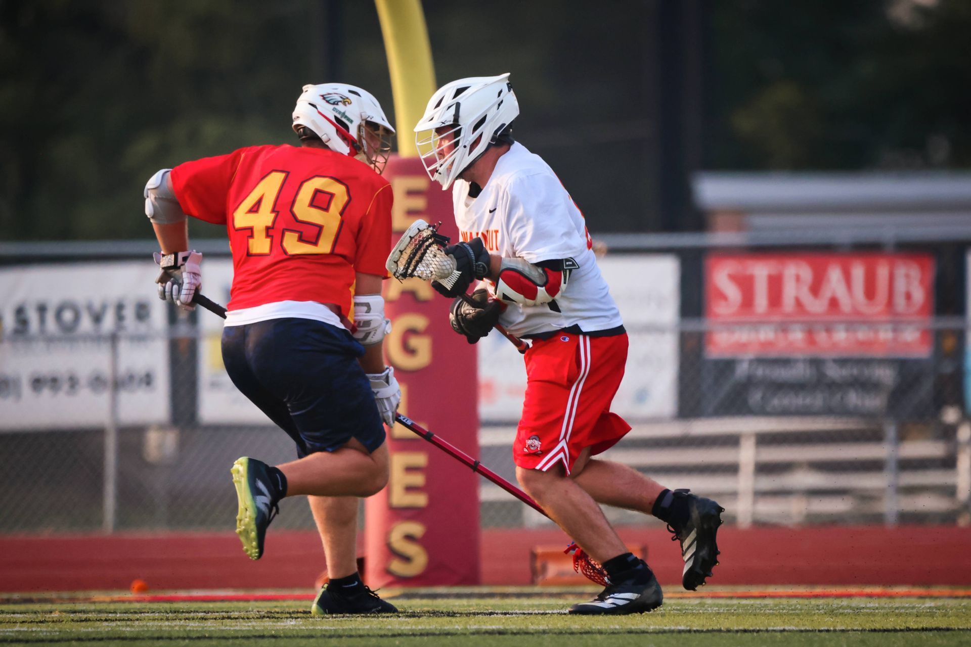 Two lacrosse players in red and white battle for the ball on a field at dusk.