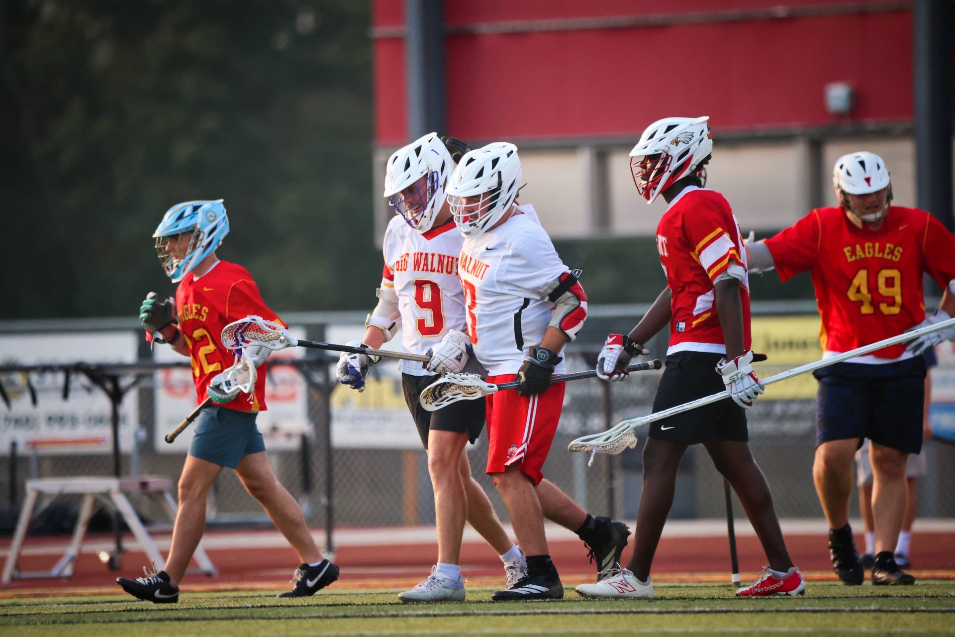 Lacrosse players in white and red uniforms stand on a field near a red wall, holding sticks.