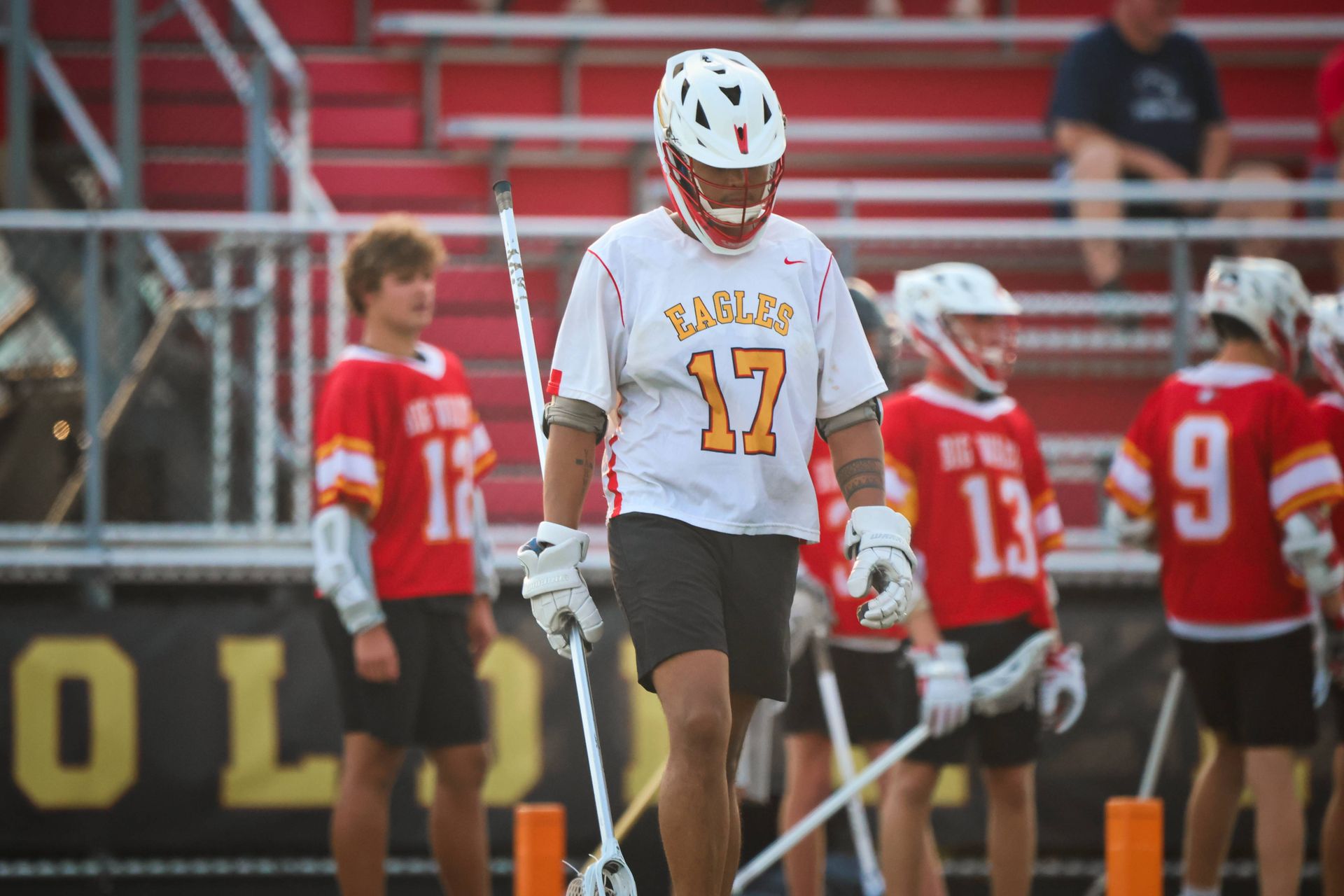 Lacrosse player in white jersey number 17 stands on the field with players in red behind him.