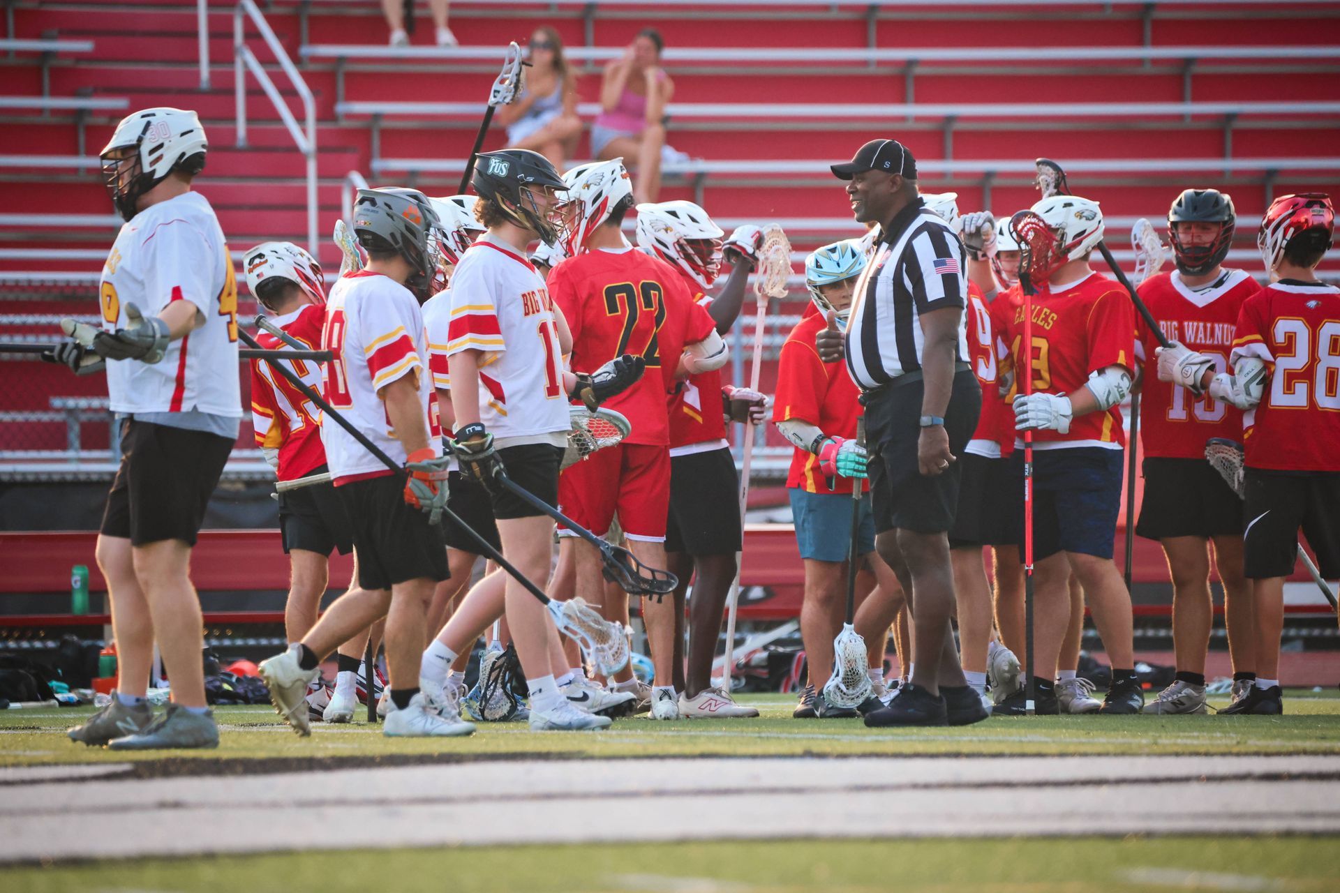 Lacrosse players and coaches gather on the sideline in red and white uniforms during a game.