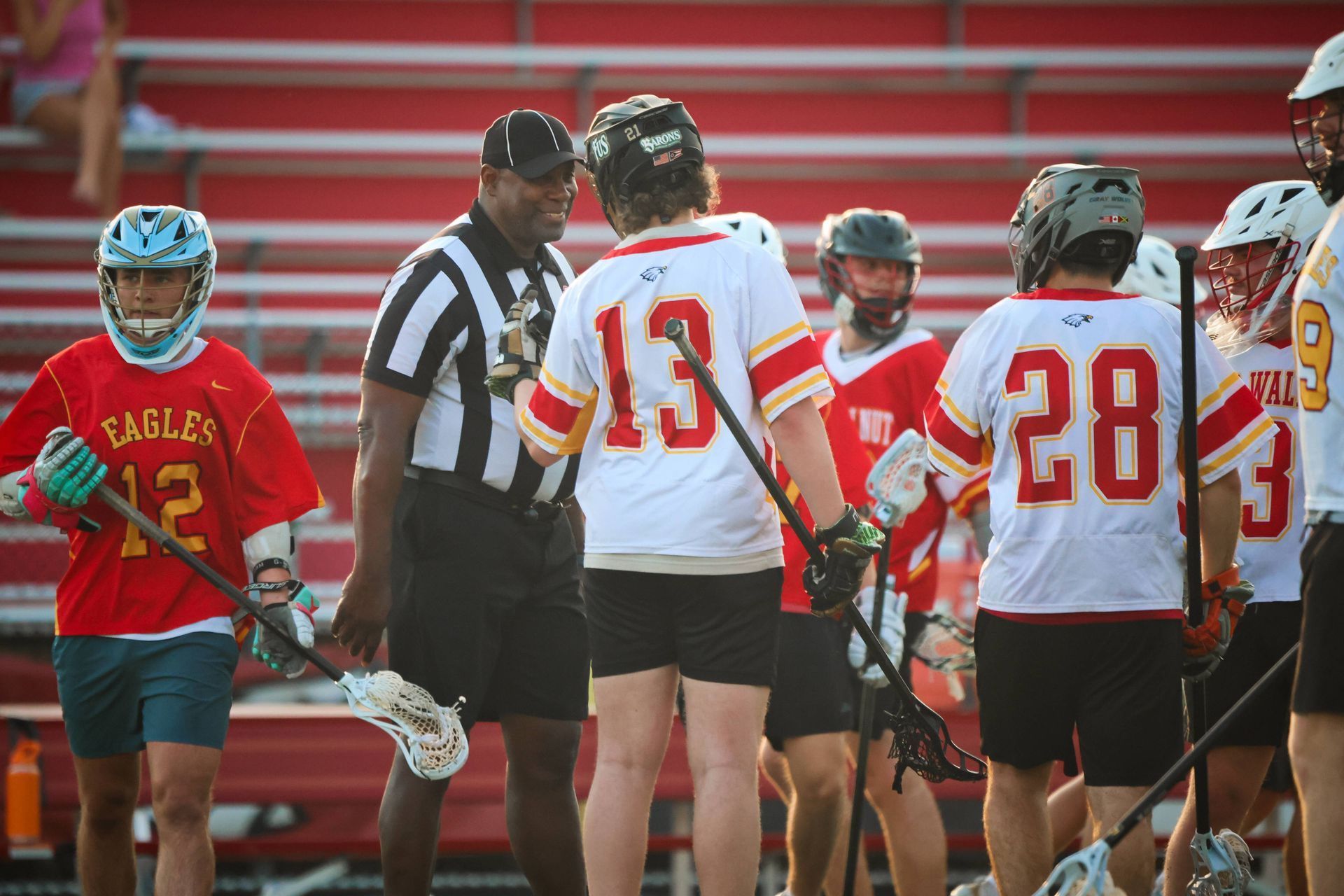 Lacrosse players huddle on the field with a coach near red bleachers, in red and white uniforms.