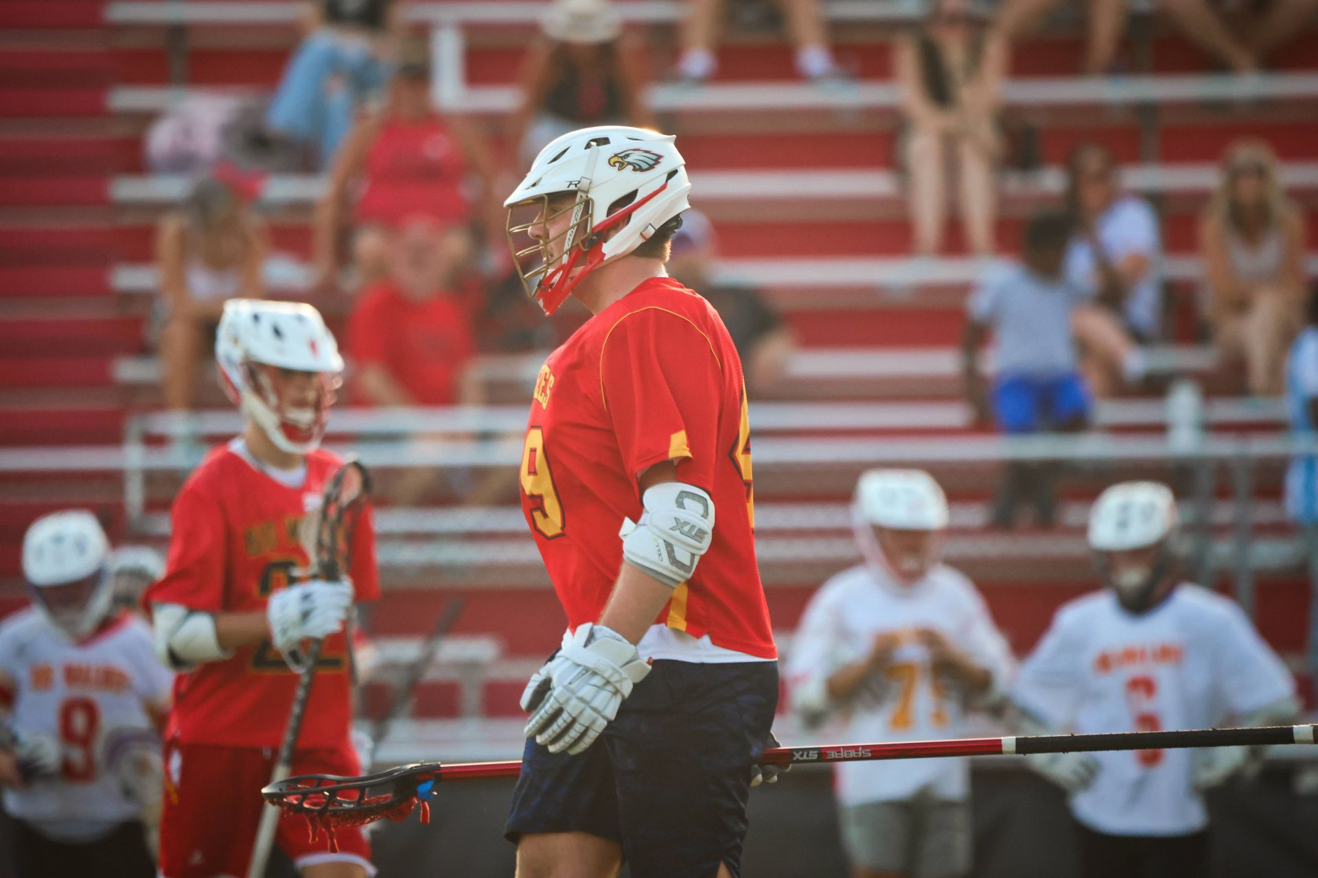 Lacrosse players in red and white jerseys stand on a field with stadium bleachers in the background