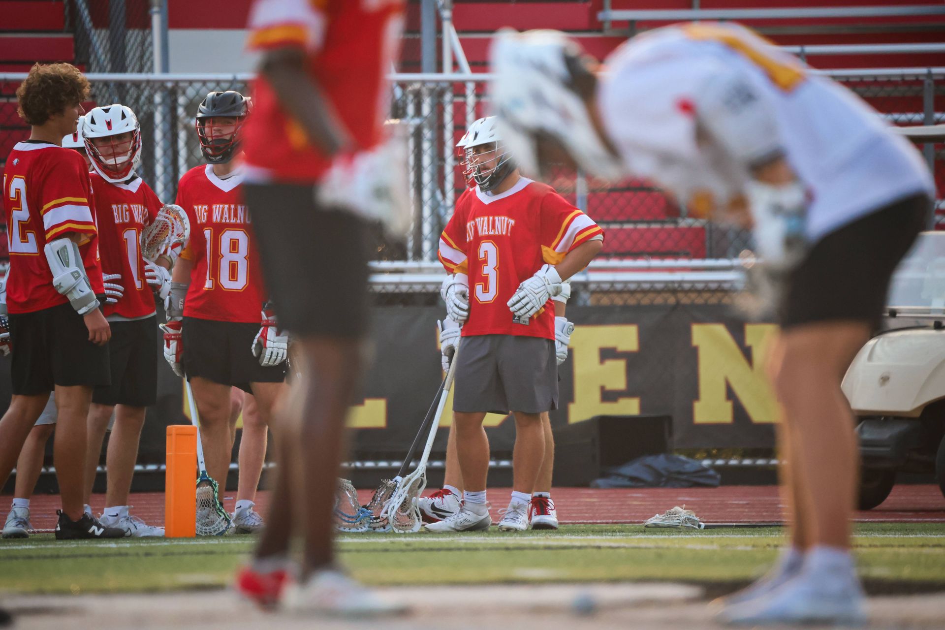 Lacrosse players in red jerseys stand on the field during a game, with blurred players in the foreground.