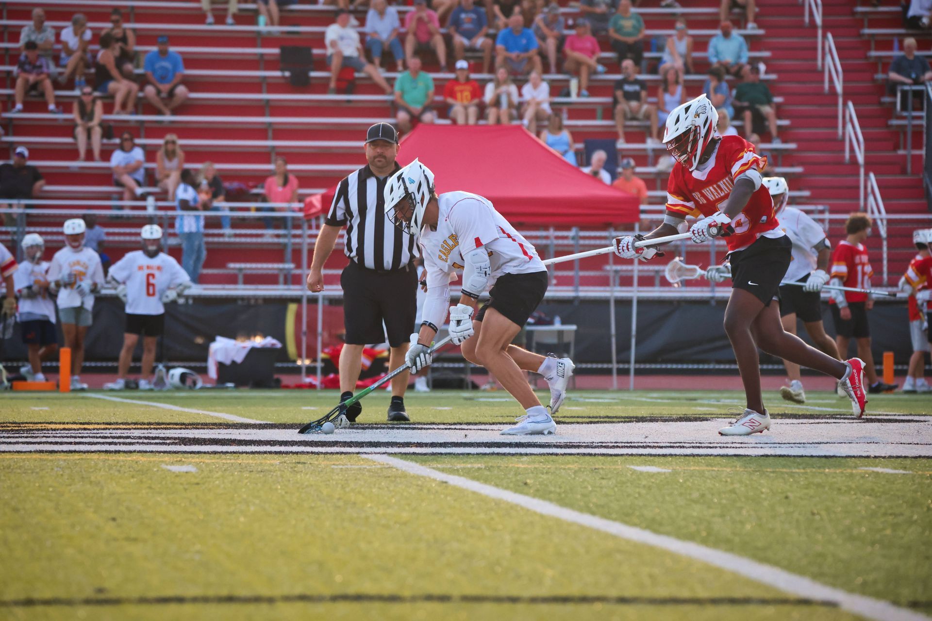 Lacrosse faceoff on a field, with players in red and white and a referee nearby, stadium seats in the background