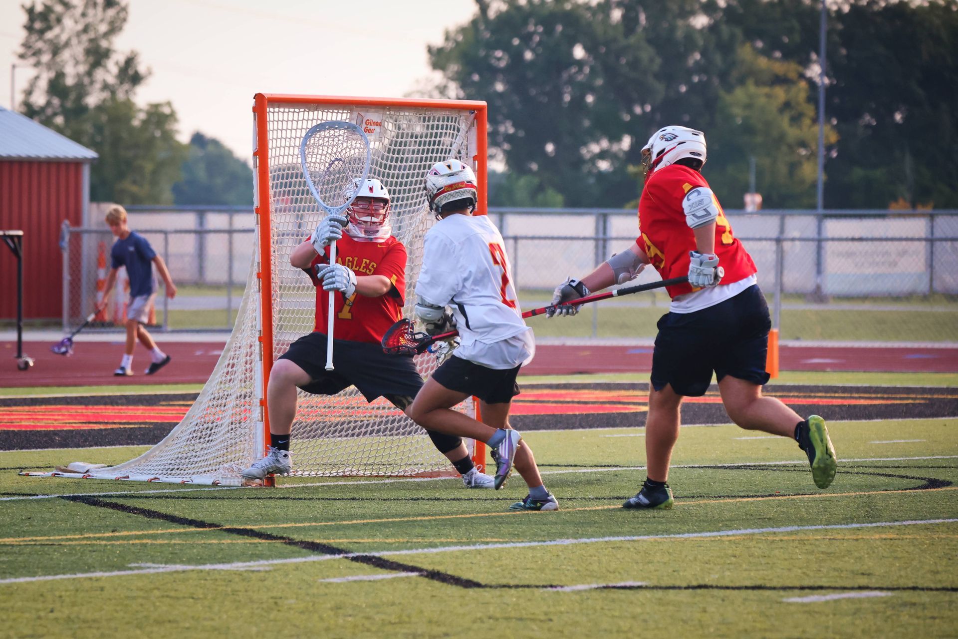 Lacrosse players in red and white battle near the goal on a field at dusk.