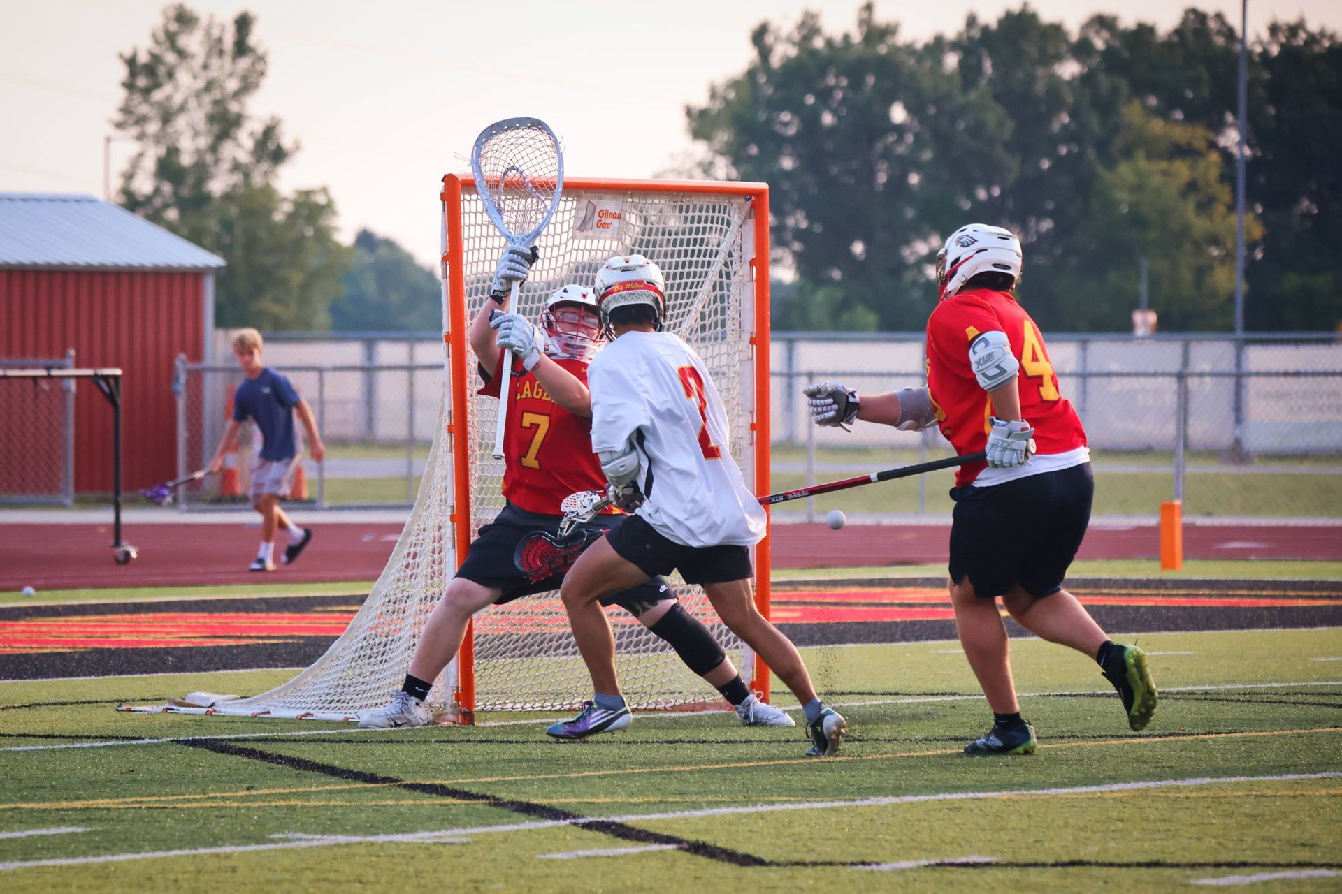 Lacrosse players in red and white jerseys contest a shot at the goal on a field.