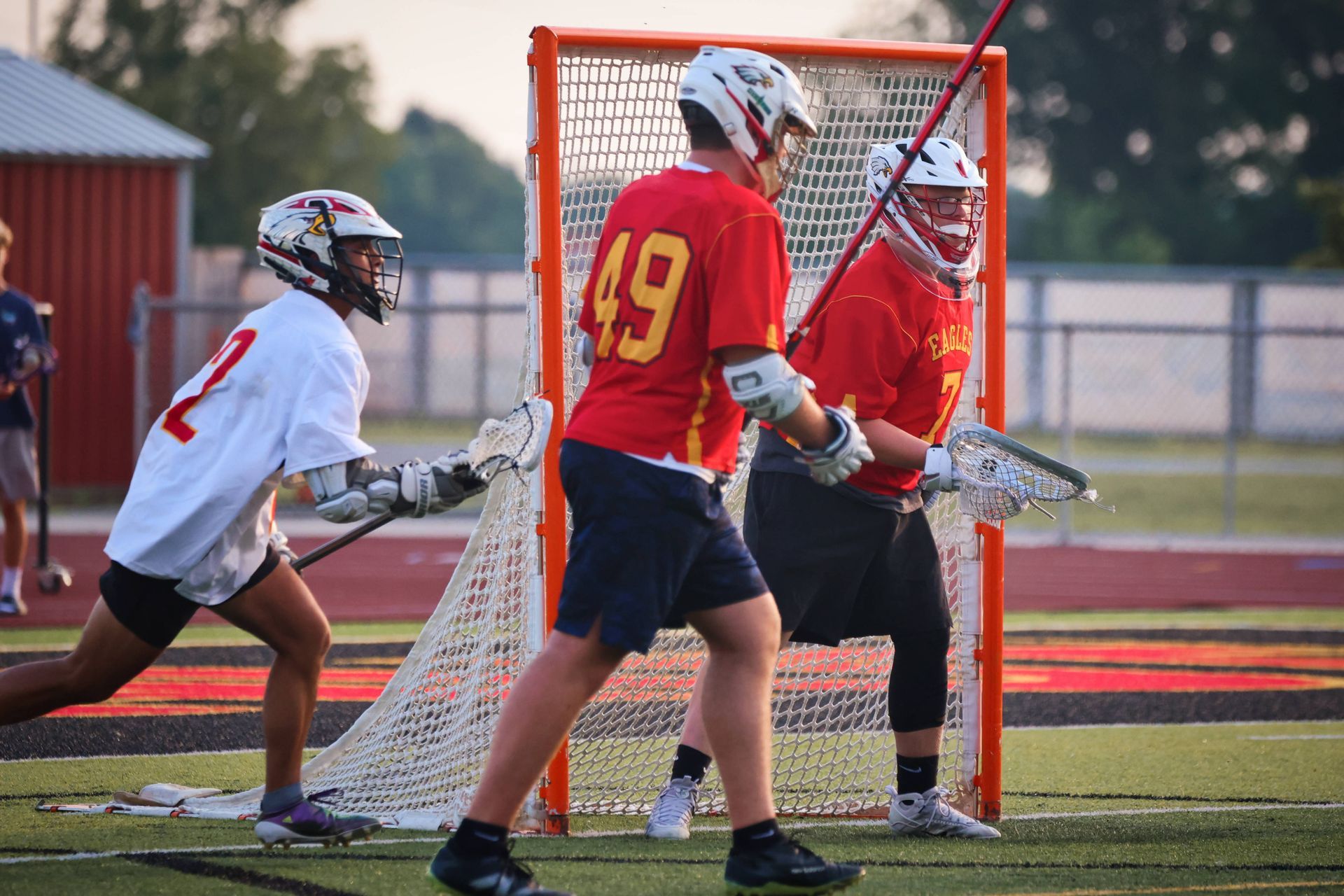 Lacrosse players in red and white uniforms contesting a shot near the goal on a field at sunset