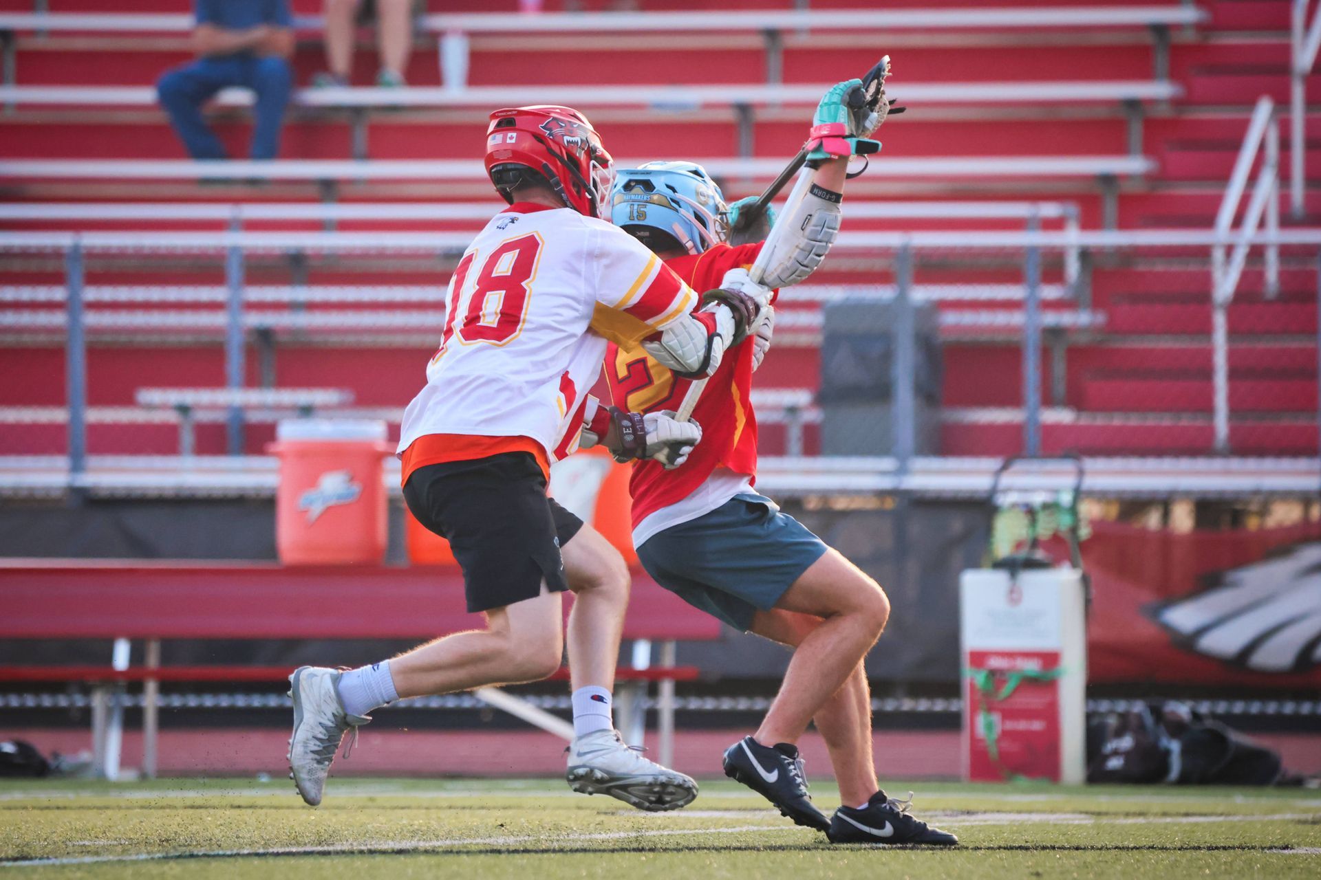 Two lacrosse players collide while contesting the ball on a field, with red stadium seats in the background.