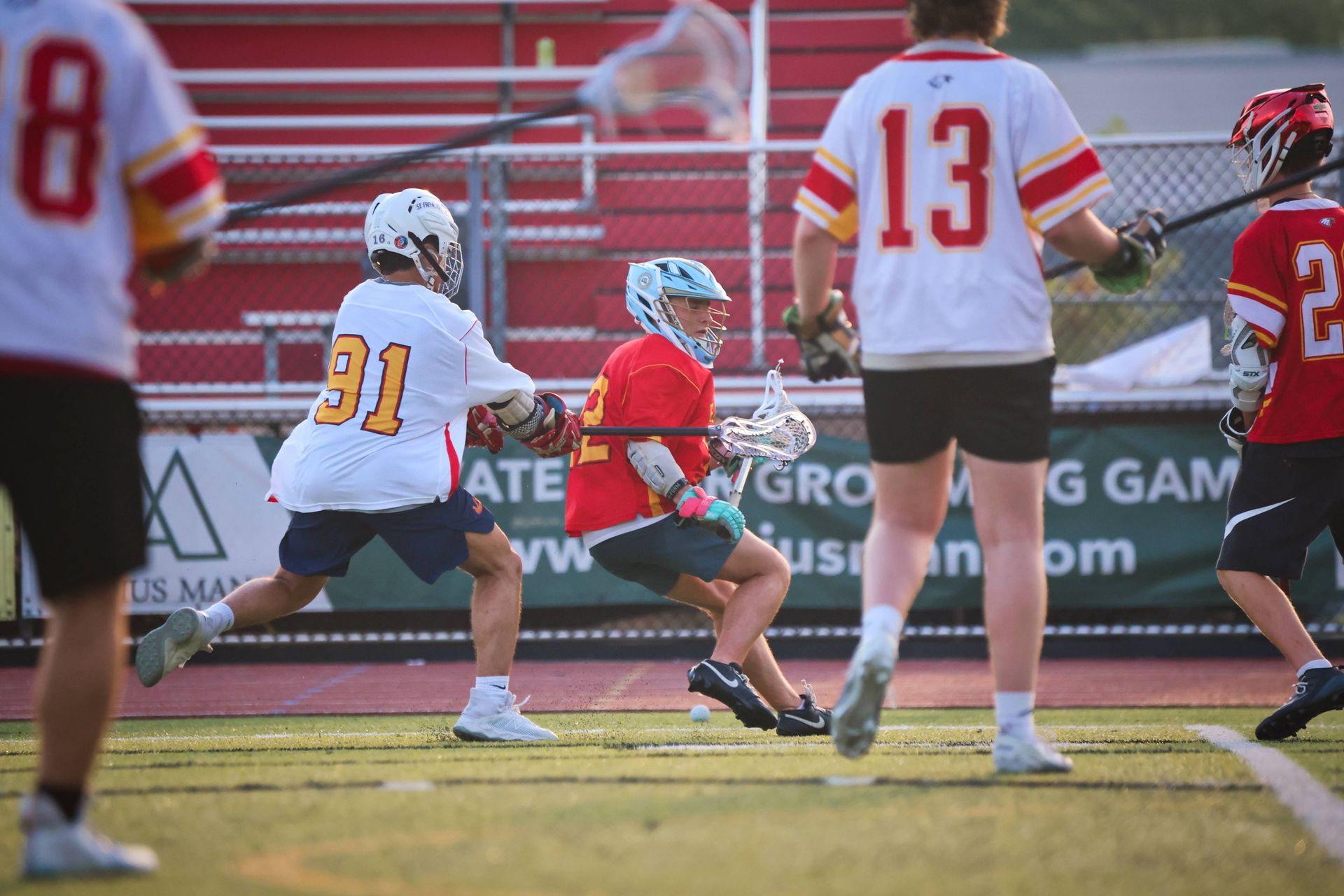 Lacrosse game with players in white, red, and black on a grassy field near the goal area
