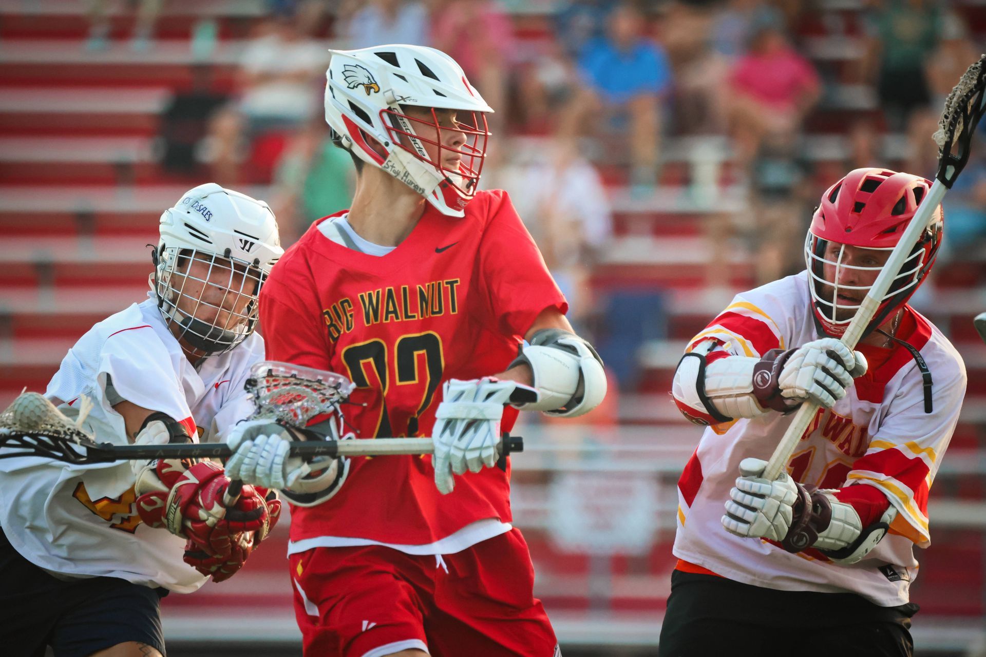 Three lacrosse players in action, one in red and two in white, on a field with blurred stands behind them