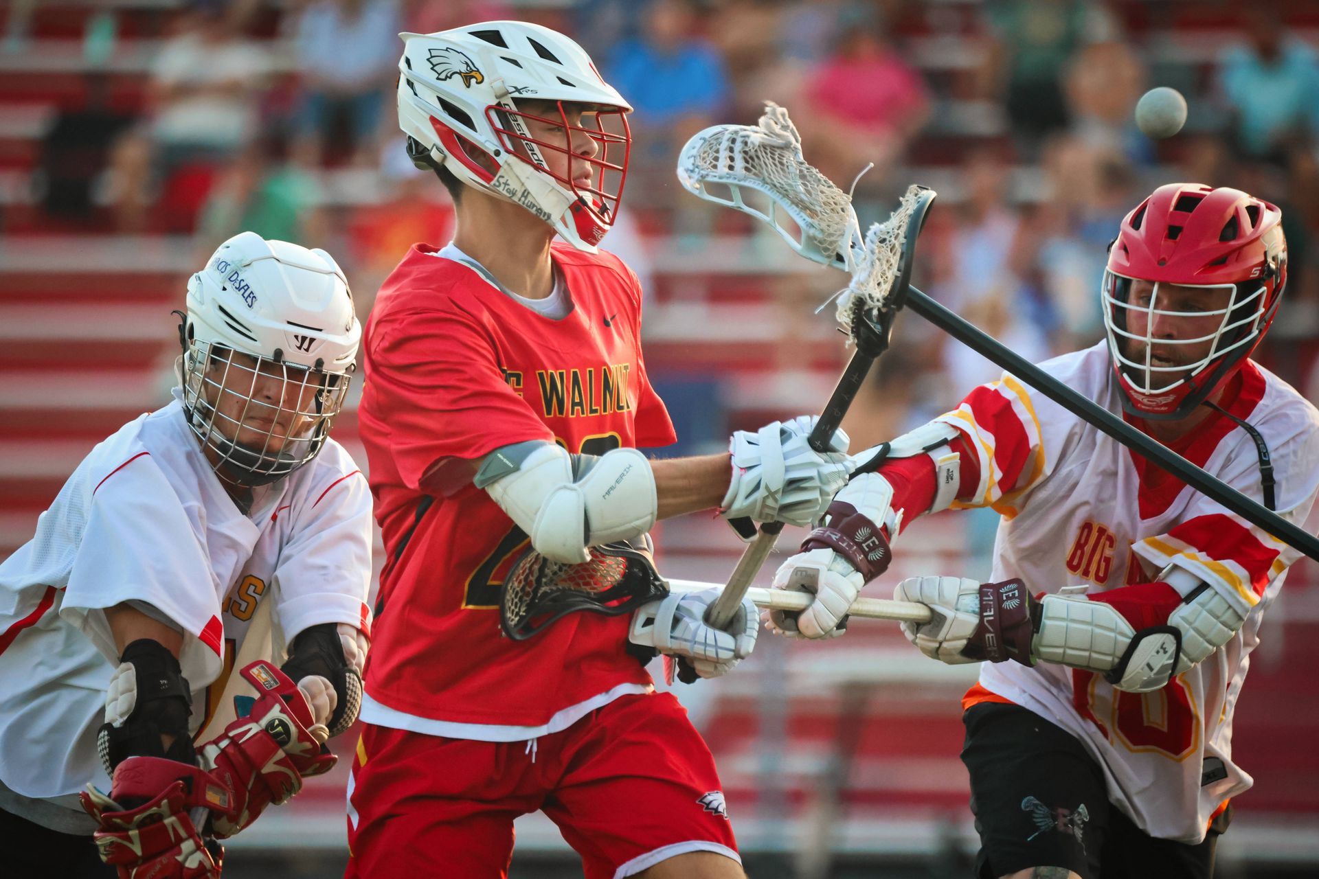 Three lacrosse players in red and white uniforms compete for the ball on a field