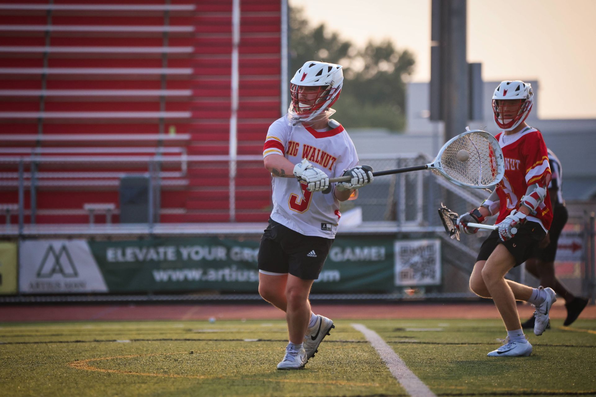 Two lacrosse players in red and white uniforms sprint on a field, one carrying the ball near the goal.