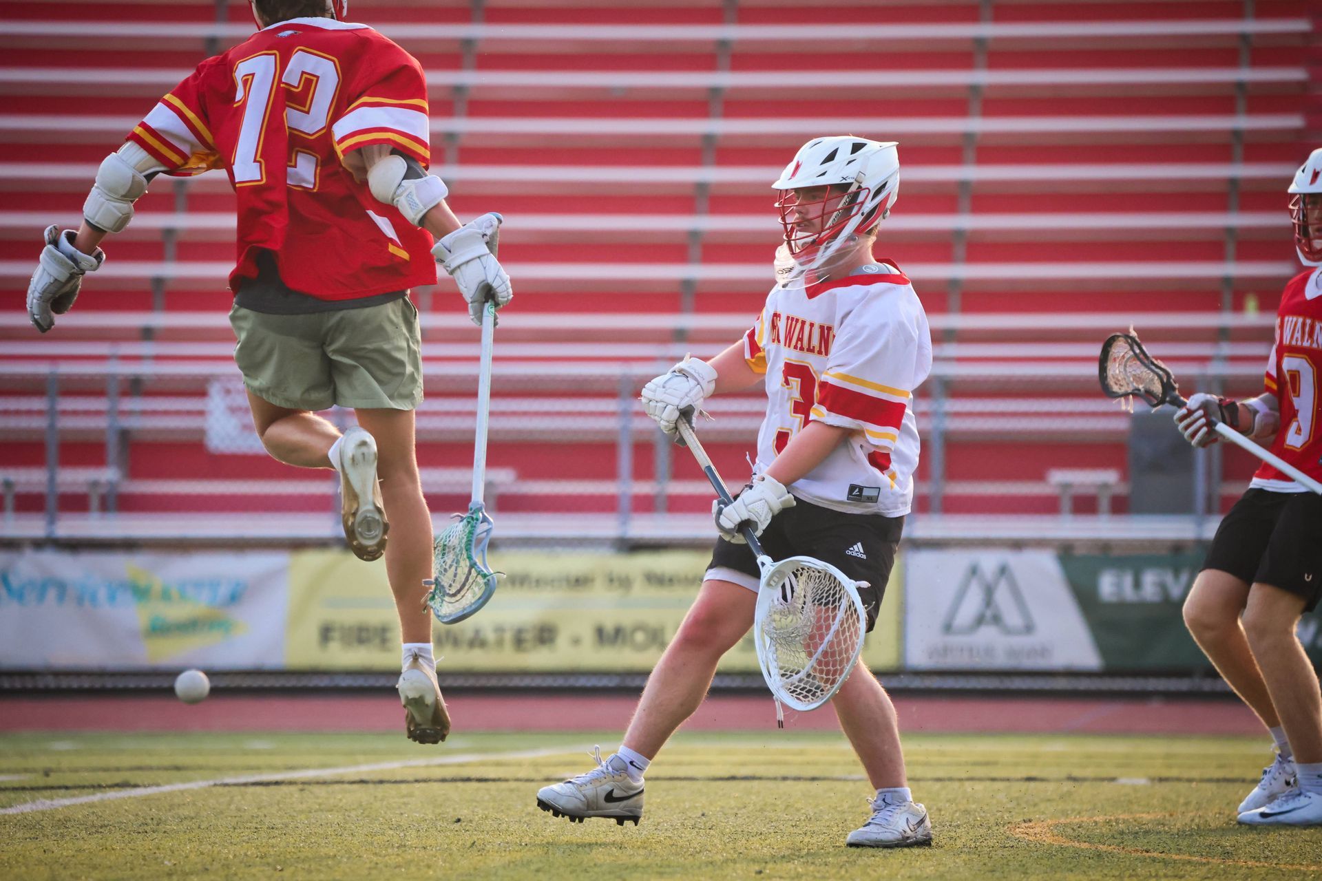 Lacrosse players in red and white uniforms running on a field in front of bleachers.