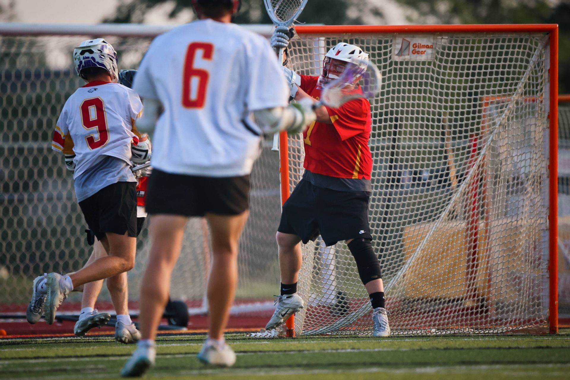 Lacrosse goalie in red jersey blocks a shot at the net while players stand nearby on the field
