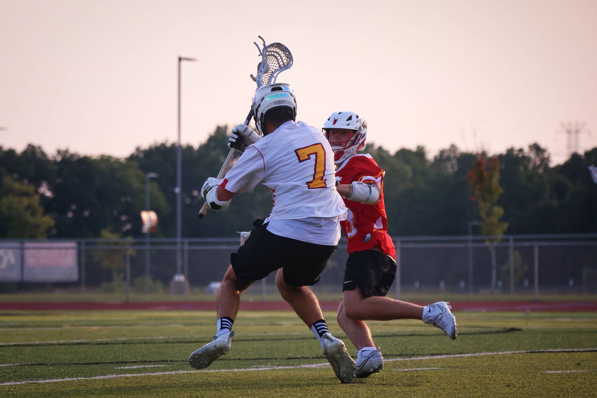 Two lacrosse players in action on a field at dusk, one in white holding the ball while being defended in red.