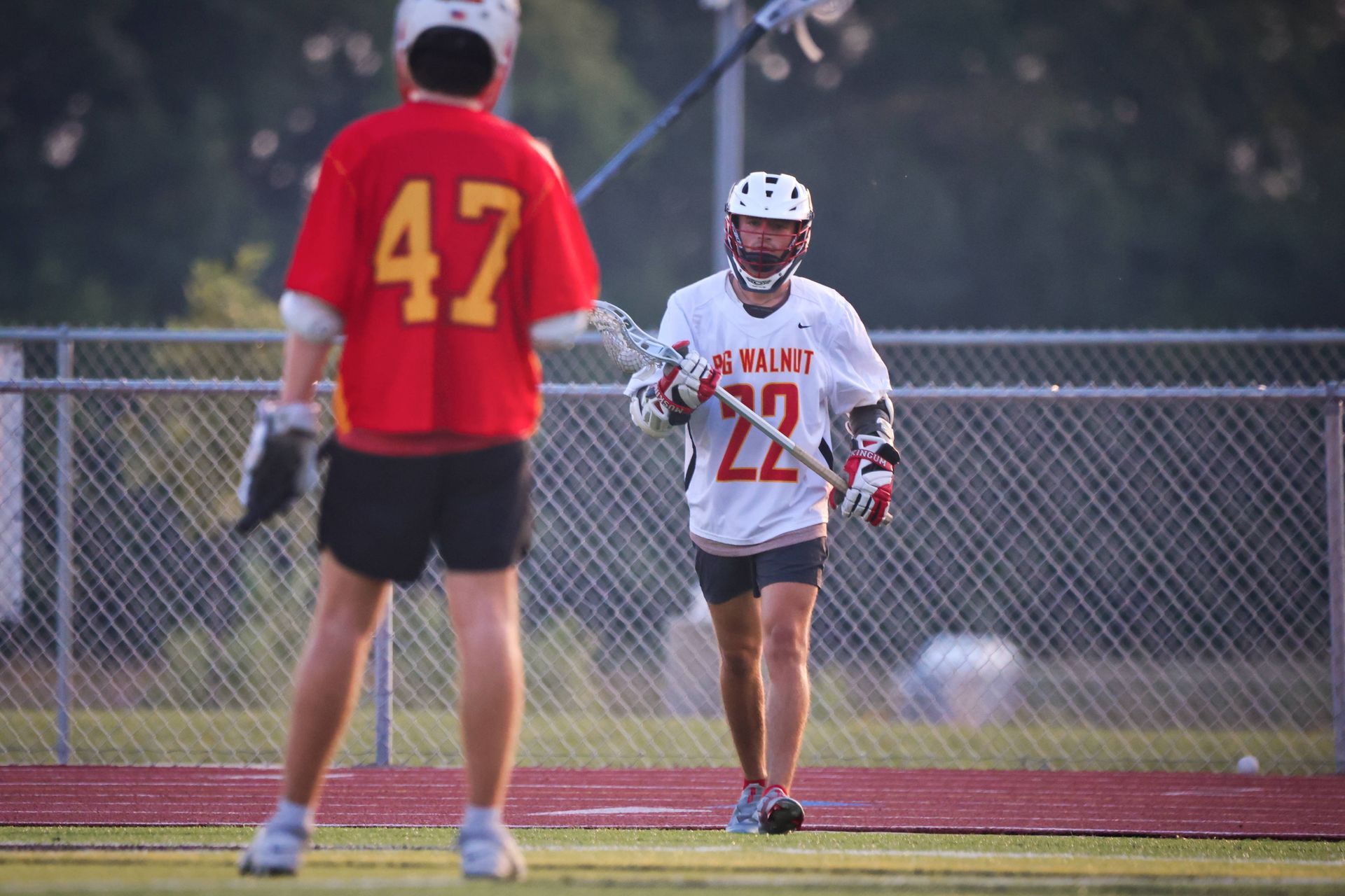 Two lacrosse players on a field, one in red jersey 47 facing another in white jersey 22 with stick raised.