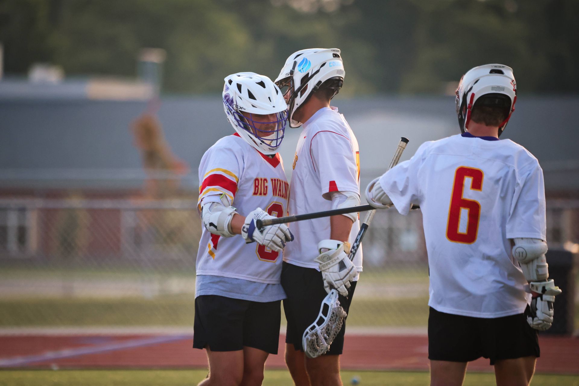 Three lacrosse players in white jerseys huddle on a field, one with number 6 facing away.