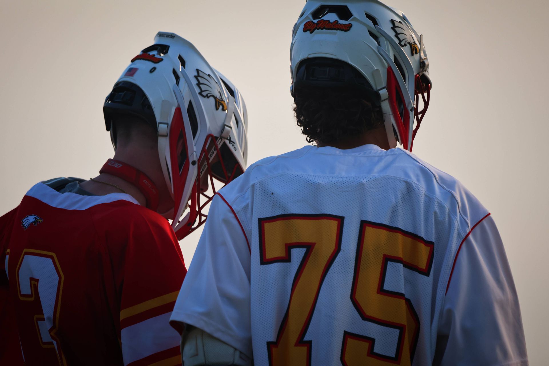 Two lacrosse players in helmets seen from behind, one in a white jersey with number 75 and one in red.