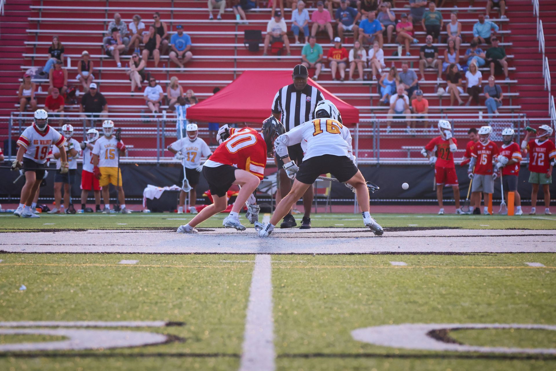 Lacrosse players lined up at the faceoff X line on a lacrosse field with red bleachers in the background