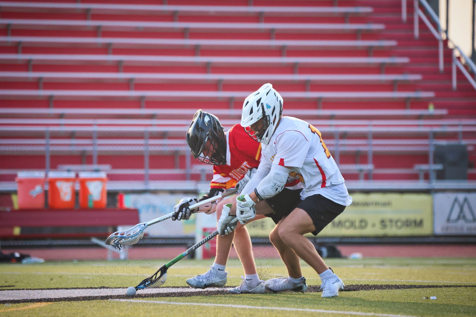 Two lacrosse players crouch for a faceoff on a field with red stadium seats behind them.