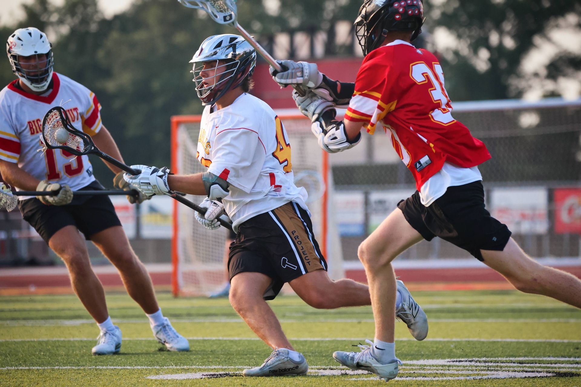 Lacrosse players in red and white uniforms compete on a field, one raising a stick to catch the ball.