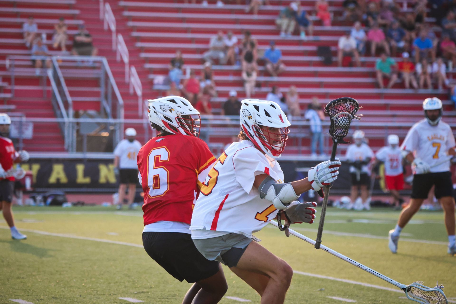 Two lacrosse players in white and red jerseys contest the ball on a field with stadium bleachers behind them.