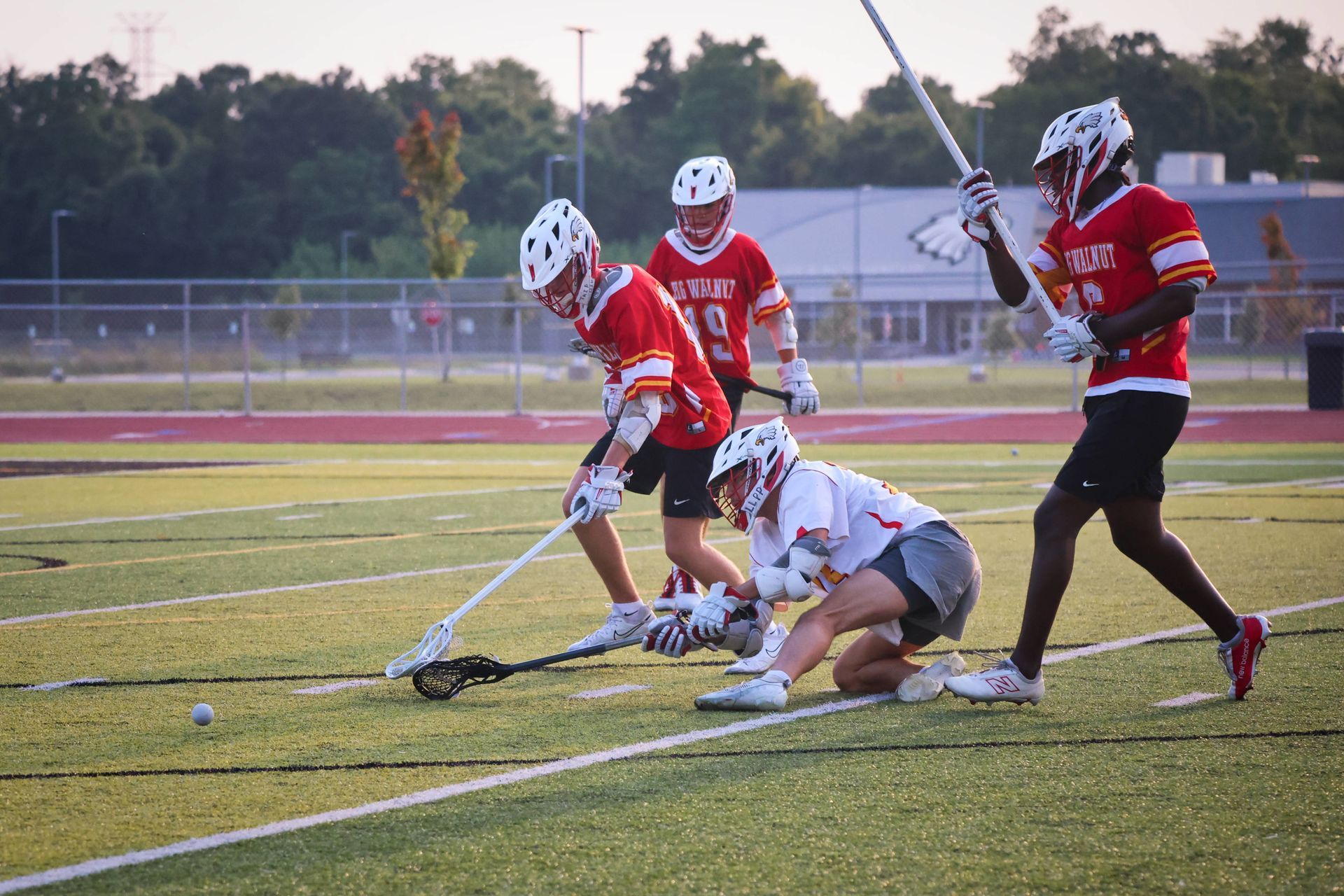 Lacrosse players in red and white battle for the ball on a field at dusk.