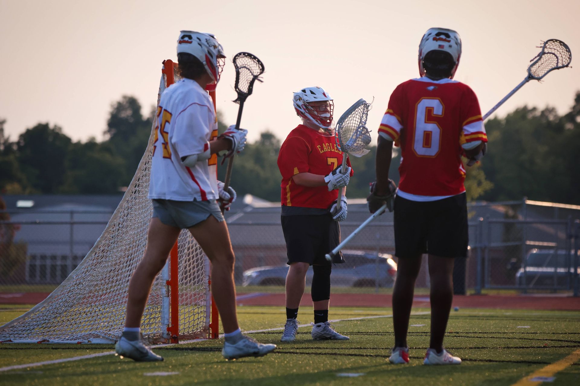 Three lacrosse players in red and white jerseys stand on a field at dusk, holding sticks near the goal.