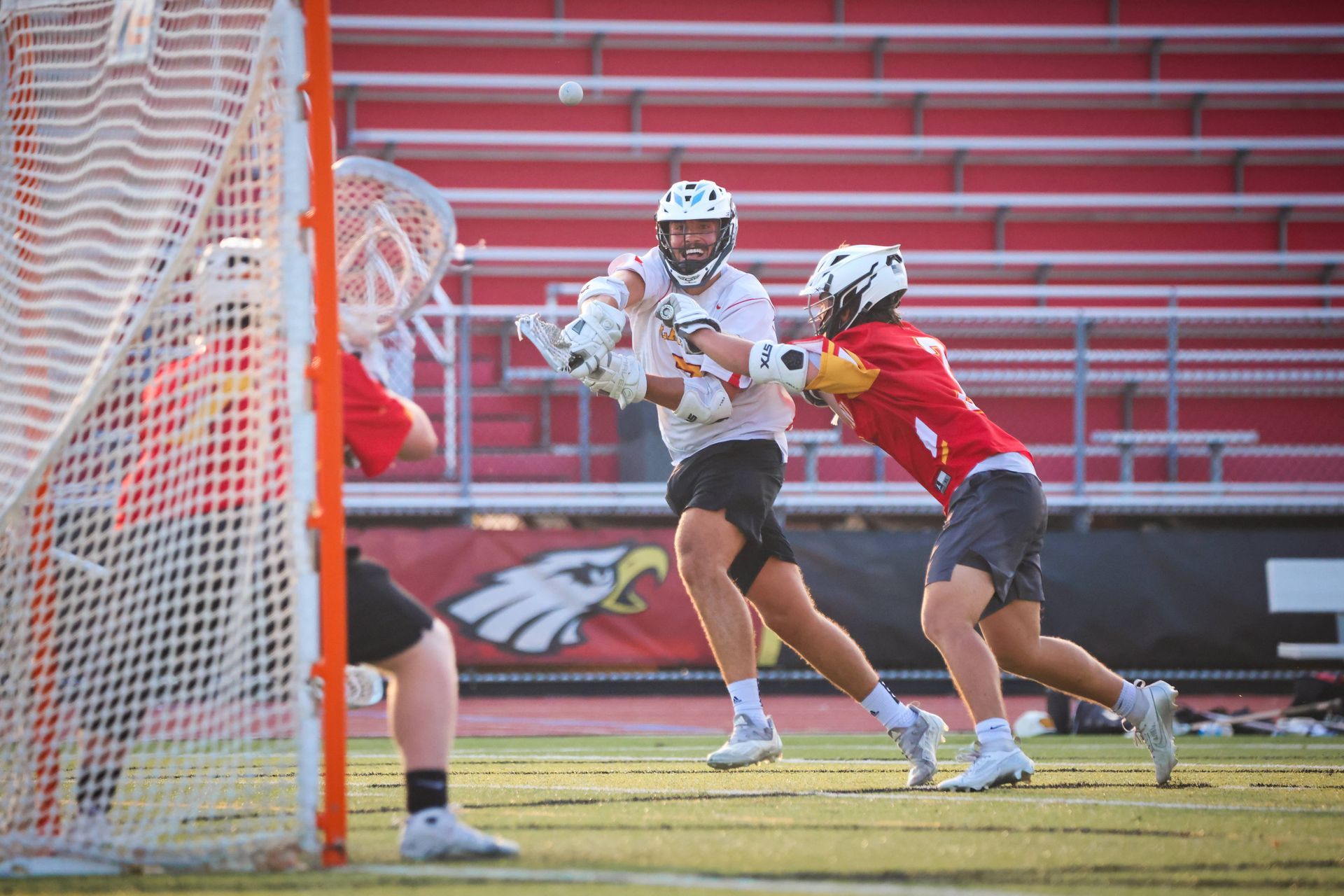 Lacrosse player in white dodges a defender in red near the goal on a field.