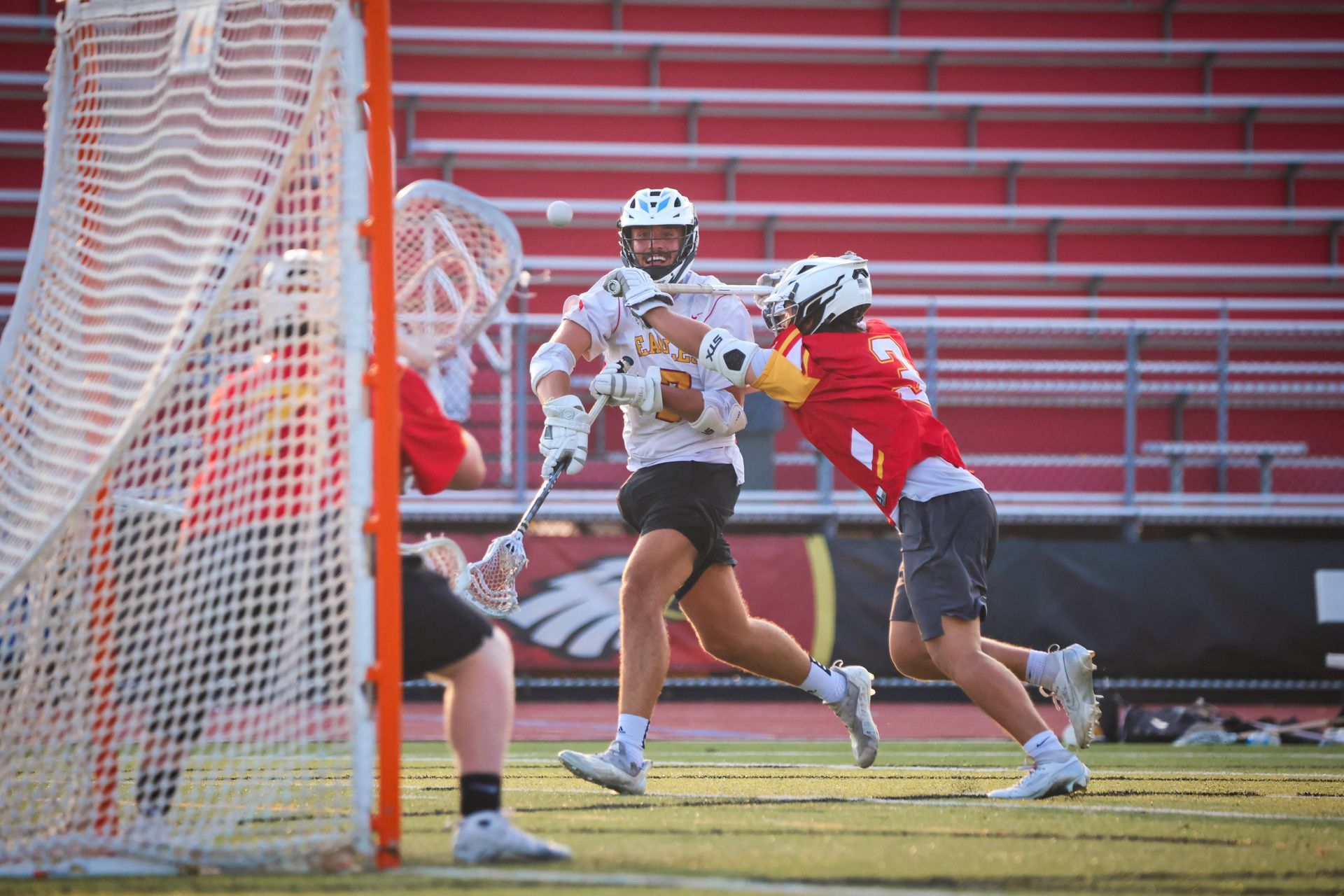 Lacrosse players in red and white contest a shot near the goal on a field with red bleachers.
