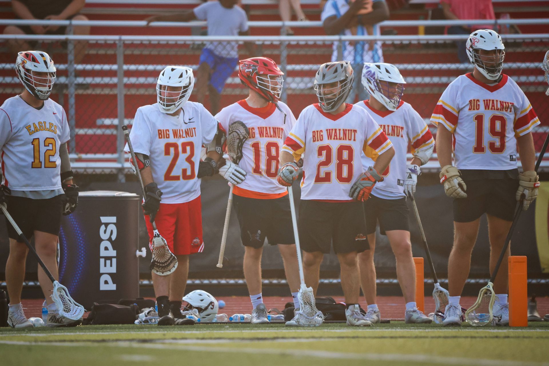 Lacrosse players in white and red uniforms stand on the sideline near the field