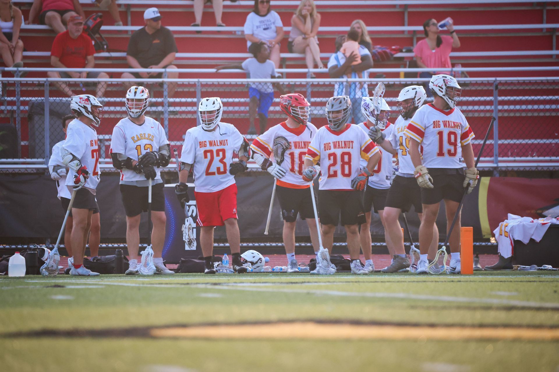 Lacrosse players in white and red uniforms line the sideline during a game, with bleachers in the background