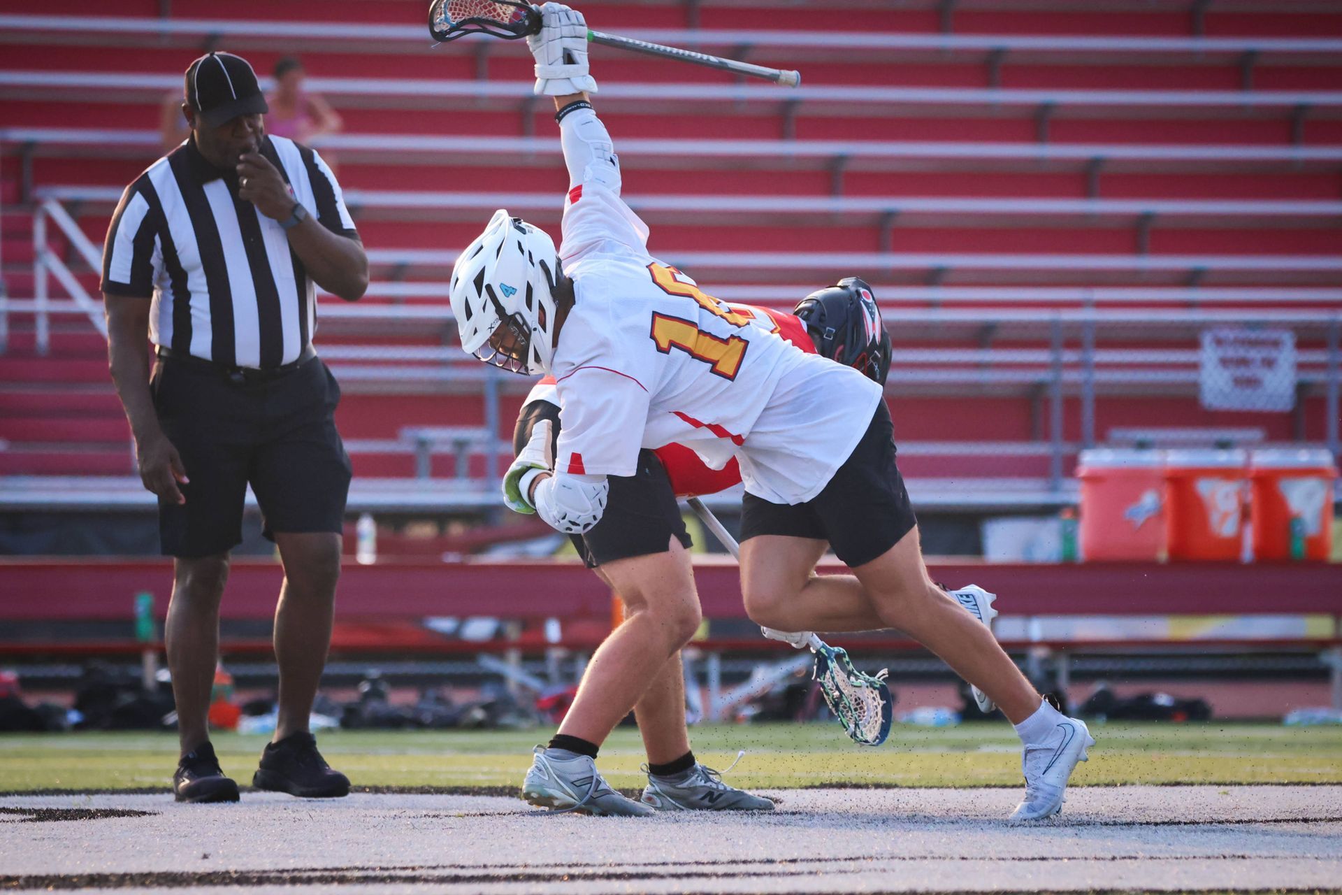 Two lacrosse players in white and red collide on the field, with a referee nearby and empty stadium seats behind them.
