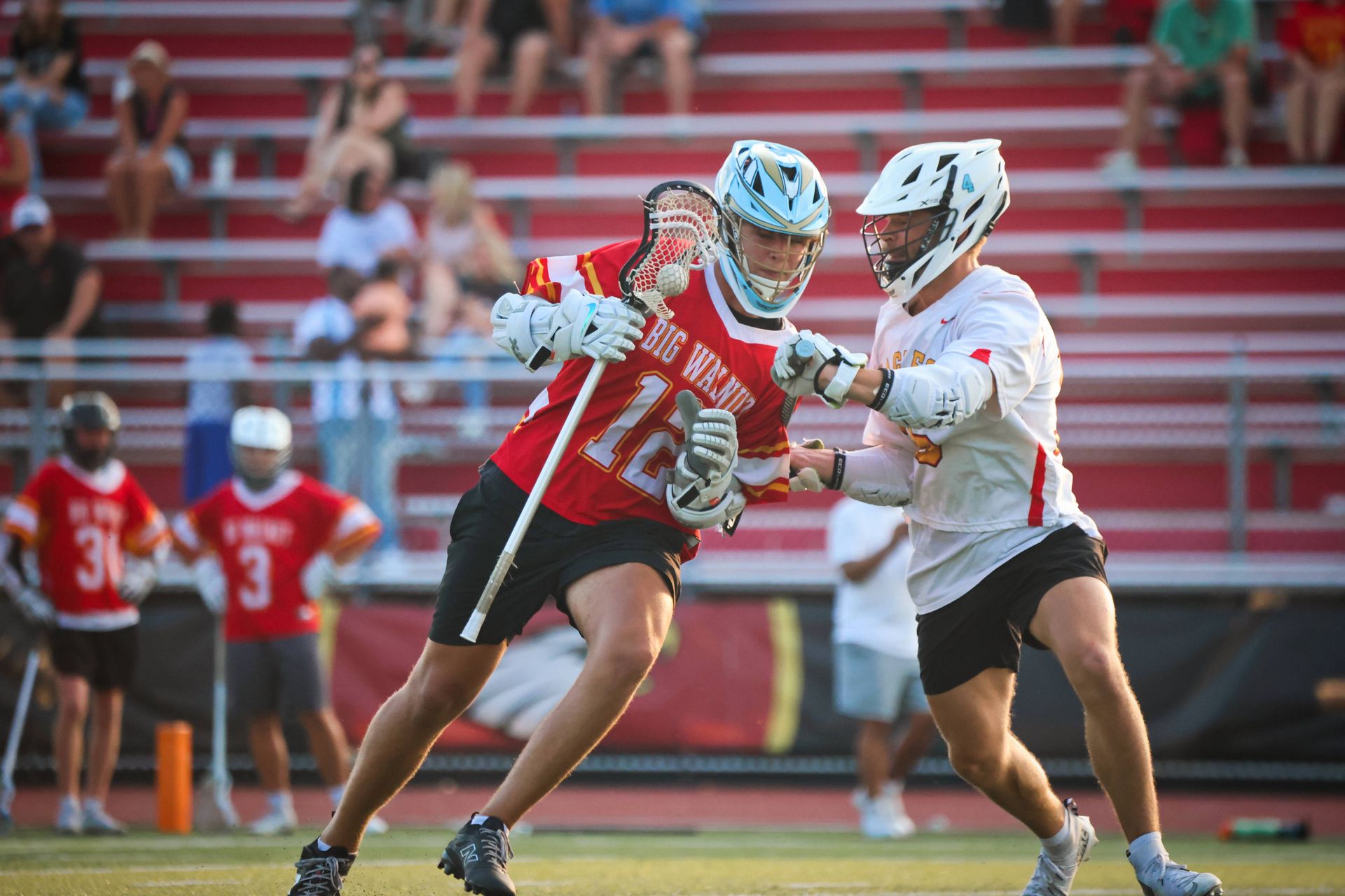 Two lacrosse players in red and white uniforms battling for the ball on a field with stadium bleachers behind them