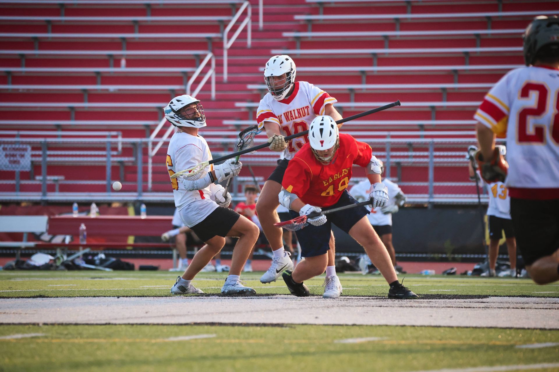Llacrosse players in red and white uniforms battle for the ball on a field with red bleachers.