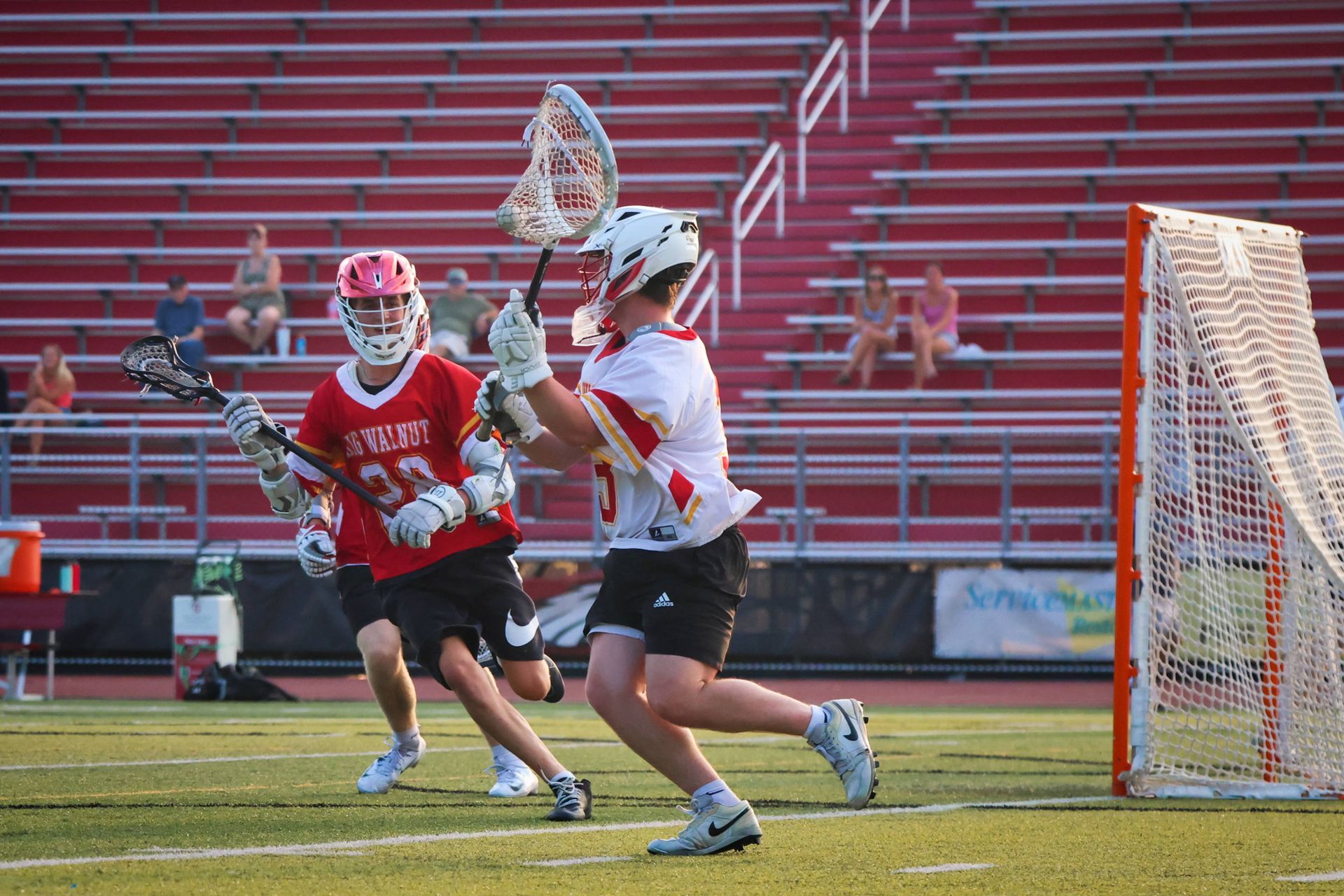 Two lacrosse players in red and white uniforms near the goal on a field