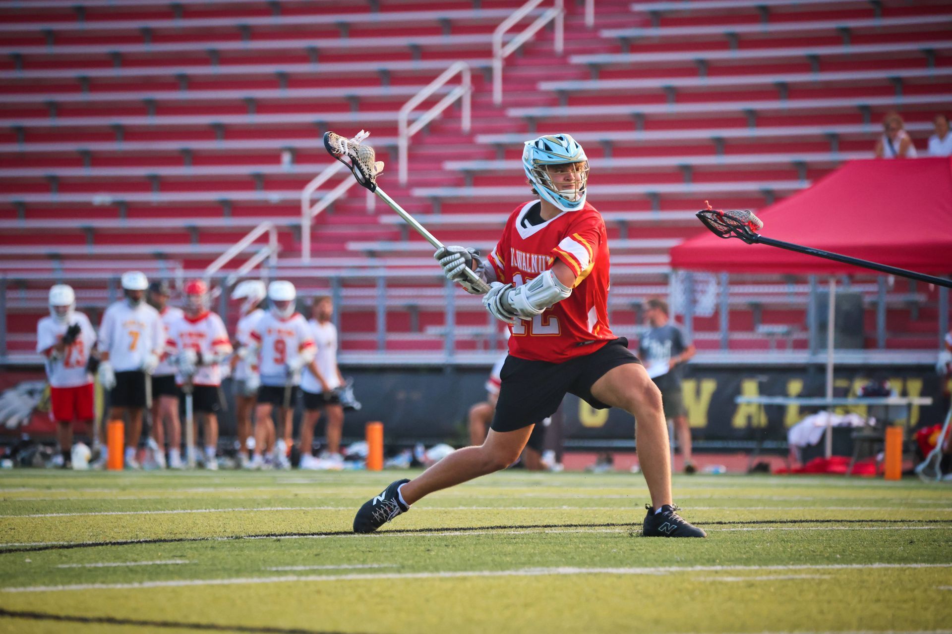 Lacrosse player in red jersey shooting the ball on a field during a game, with teammates and red bleachers behind.