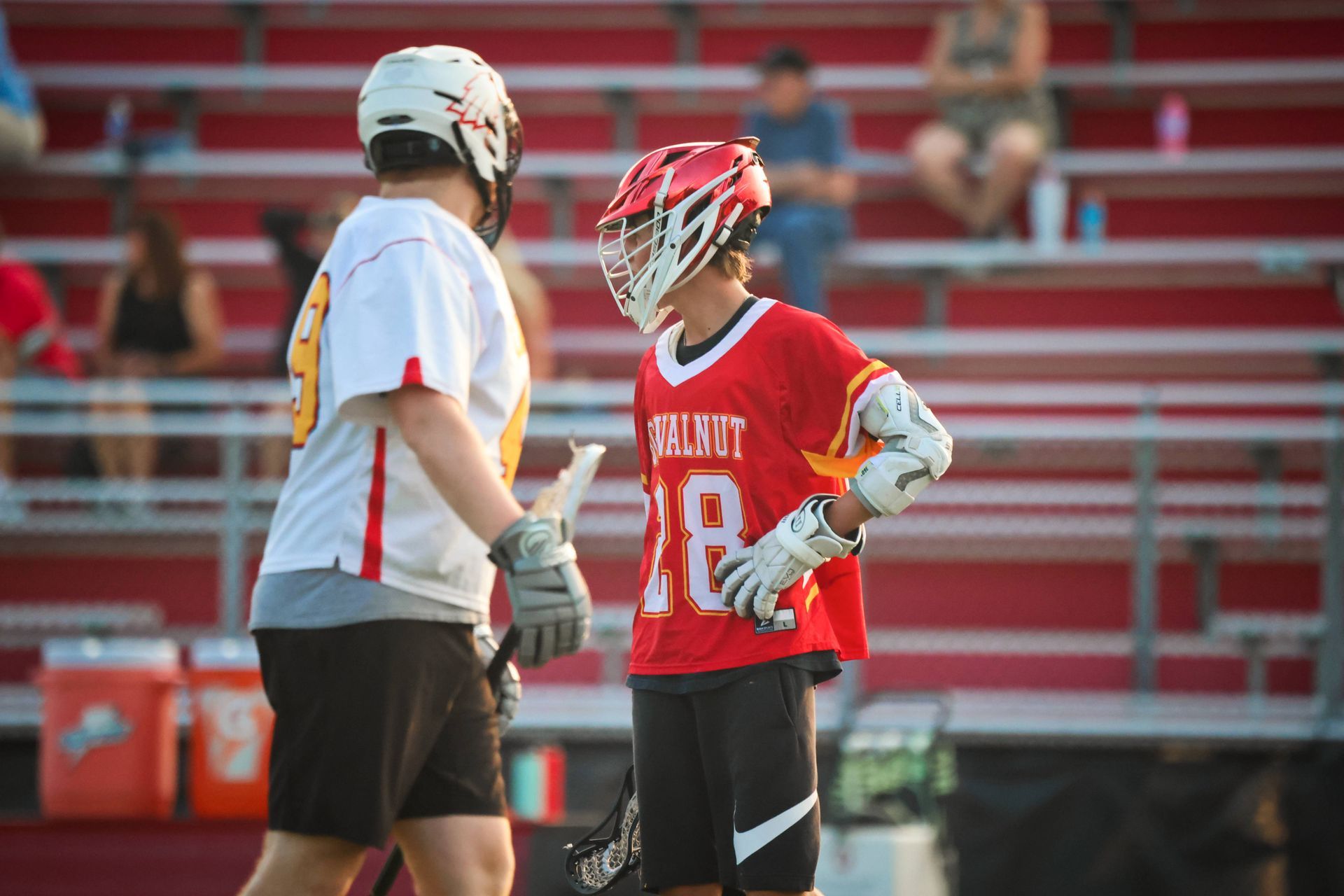 Two lacrosse players facing each other on the field, one in red and one in white, with stadium seats behind them.