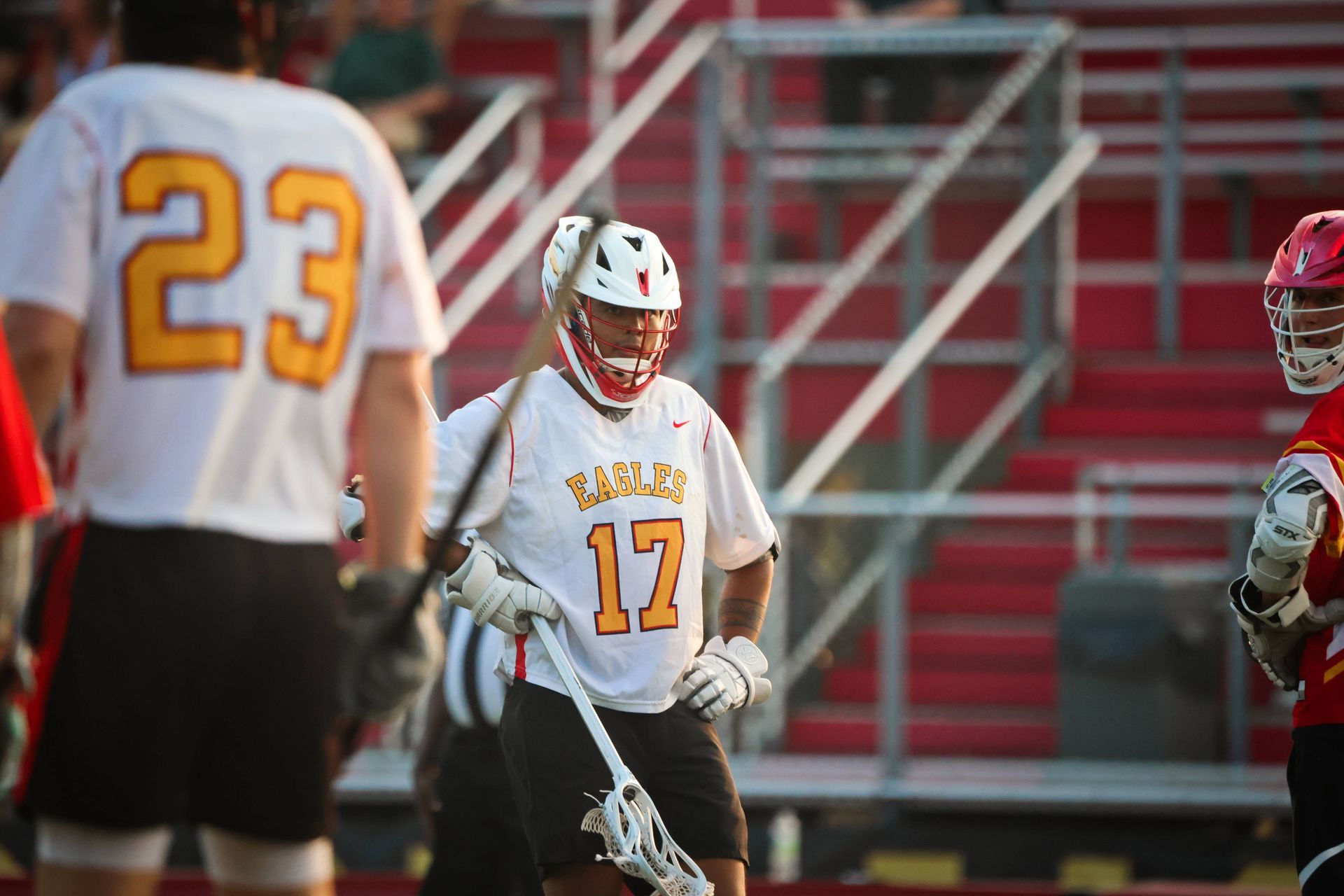 Lacrosse players in white and red uniforms on a field, with one player holding a stick near the goal.