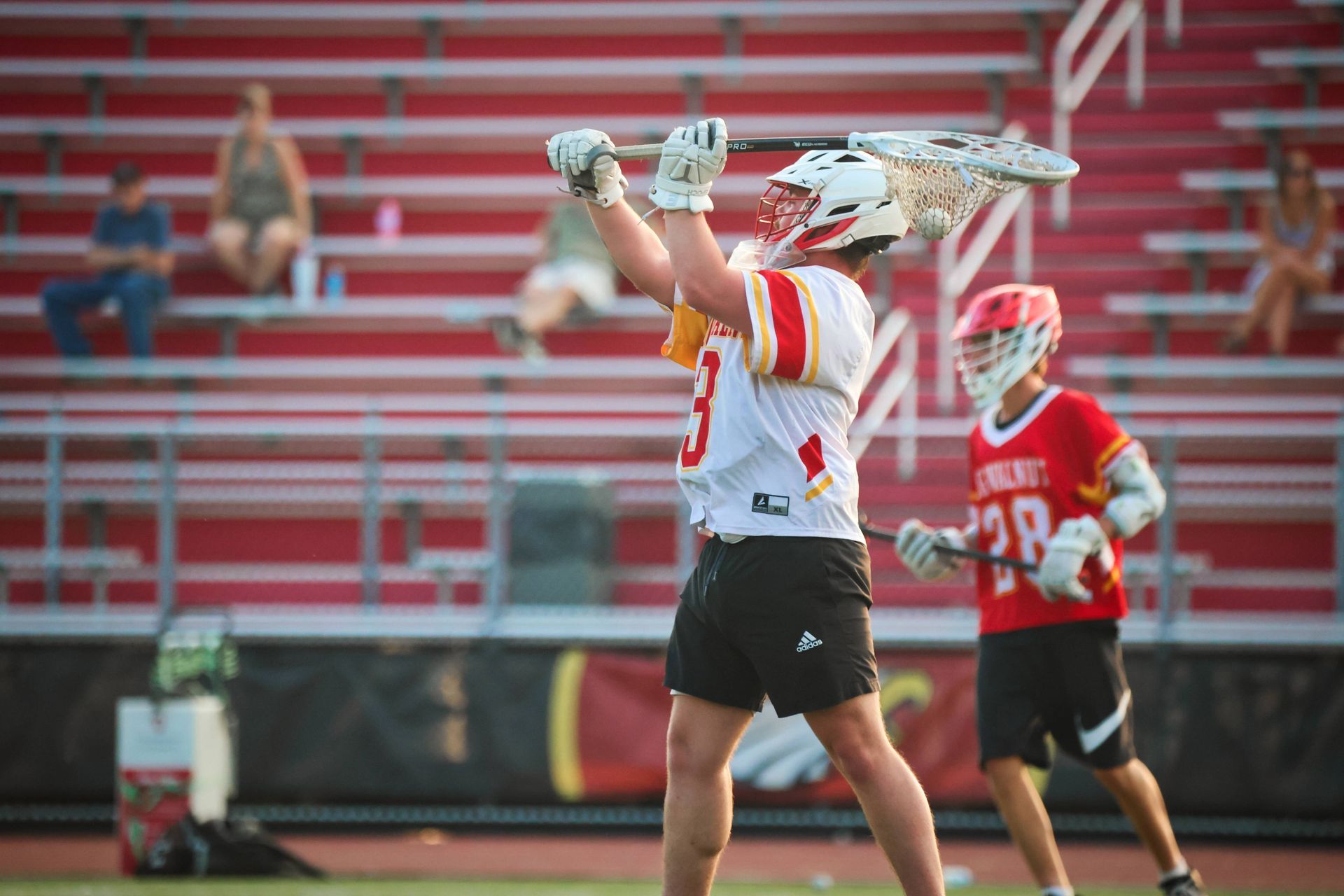 Lacrosse goalie in white raises stick to throw a clearing pass on a field, with another player and bleachers behind.