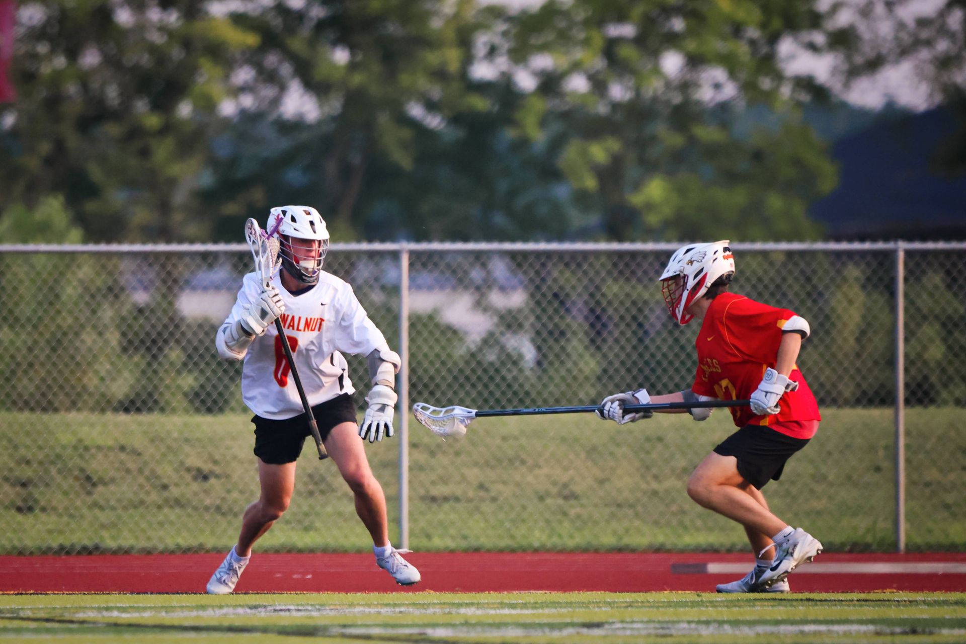 Two lacrosse players in white and red jerseys compete on a field, one defending with a stick.